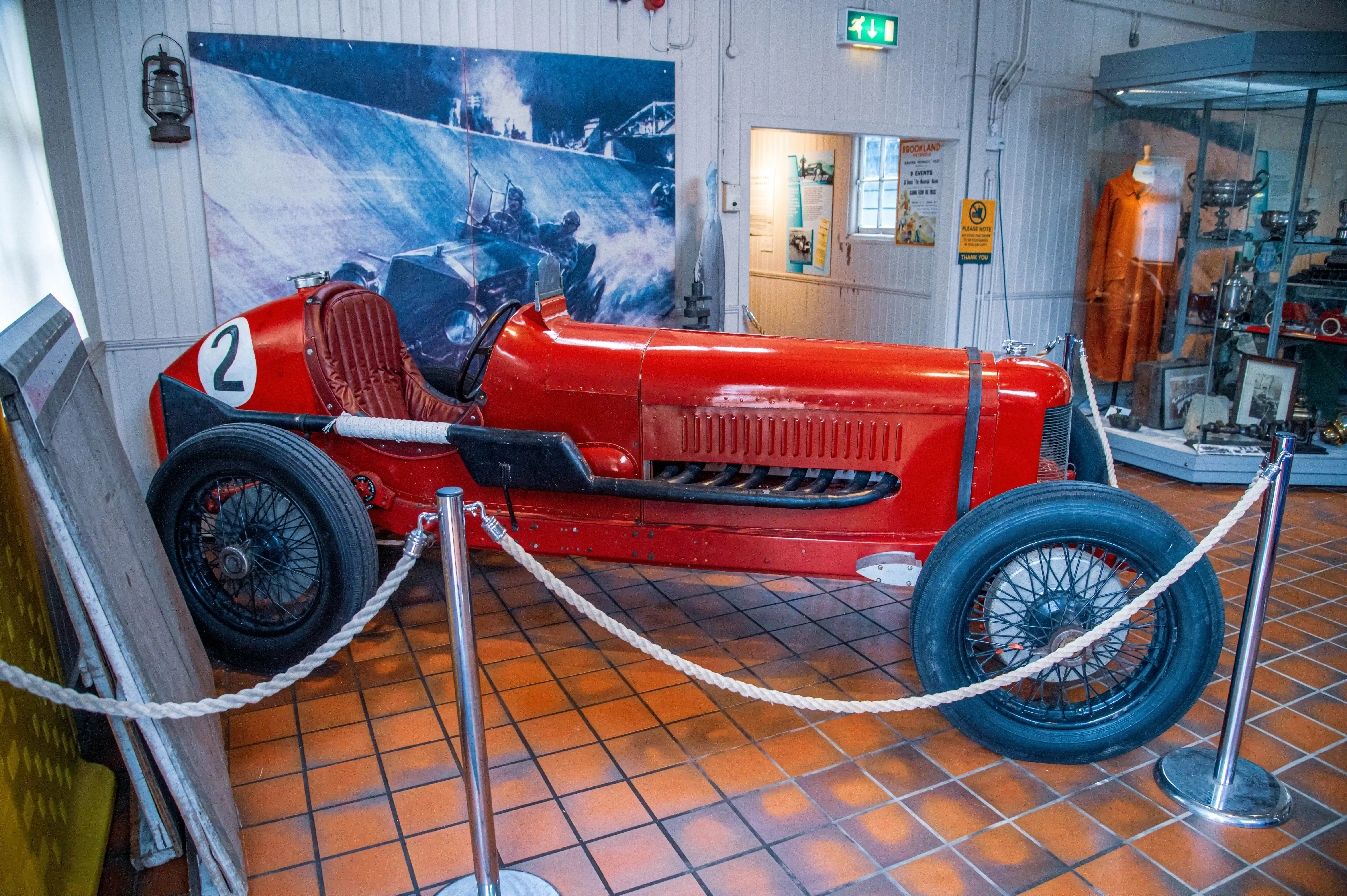 A vibrant red, vintage racing car with the number '2' displayed on its front is positioned on a terracotta tiled floor. The car has a classic design with a large grille and spoked wheels. Behind the car, a large, colourful mural shows a historic motor race with multiple cars and drivers. To the right of the mural, a life-sized cardboard cutout of a man wearing a cap and jumper stands next to an information board. The car is roped off with polished metal stanchions and thick, pale ropes.