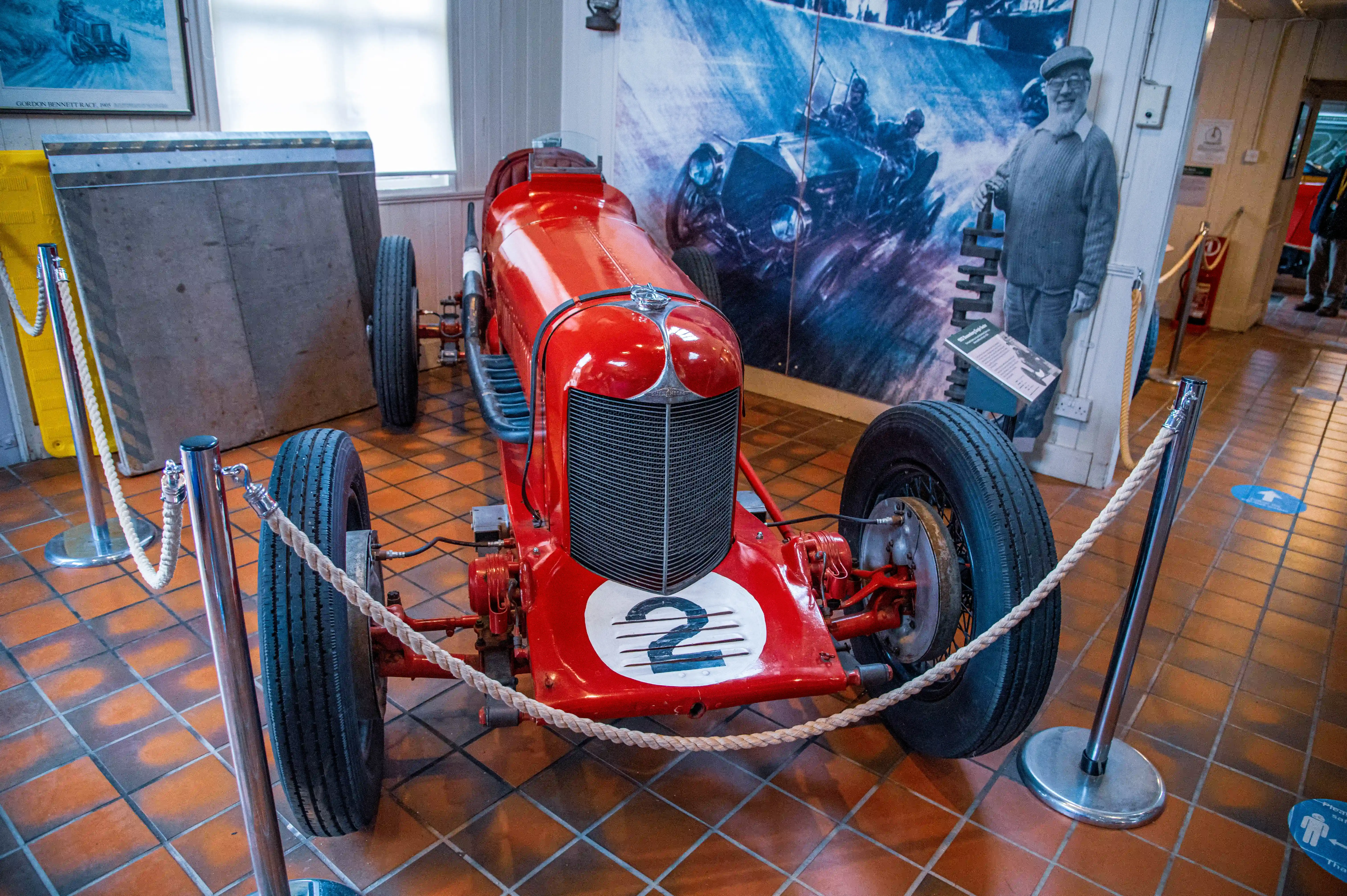A vibrant red, vintage racing car with the number '2' displayed on its front is positioned on a terracotta tiled floor. The car has a classic design with a large grille and spoked wheels. Behind the car, a large, colourful mural shows a historic motor race with multiple cars and drivers. To the right of the mural, a life-sized cardboard cutout of a man wearing a cap and jumper stands next to an information board. The car is roped off with polished metal stanchions and thick, pale ropes.