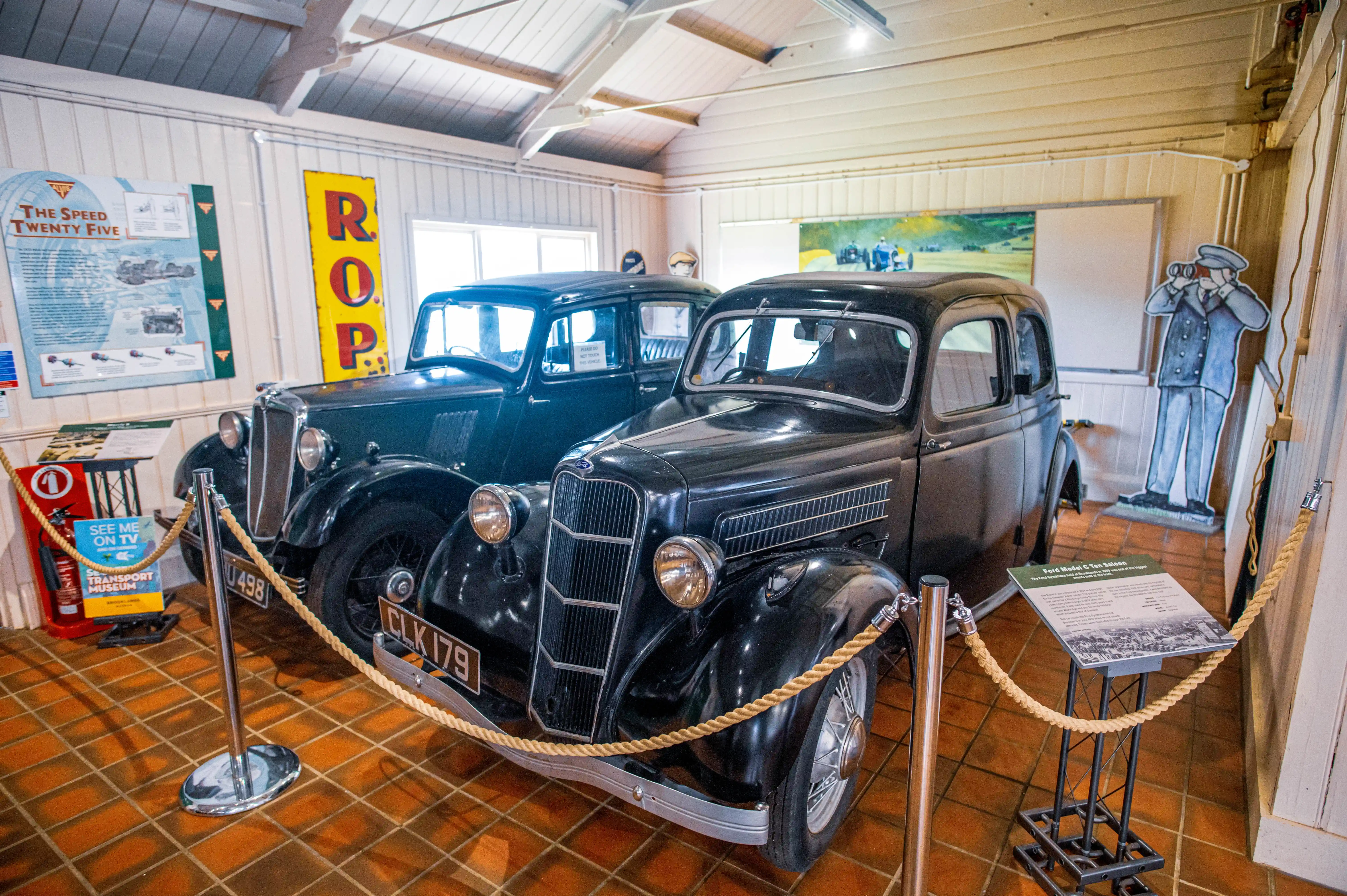 Two vintage cars, a dark green one and a black one, are parked side-by-side in a museum setting. Rope barriers separate the viewer from the vehicles. Informative signs and posters are visible on the walls, along with a life-sized cardboard cutout of a man in a uniform holding binoculars. The cars have classic styling with prominent grilles and rounded shapes.