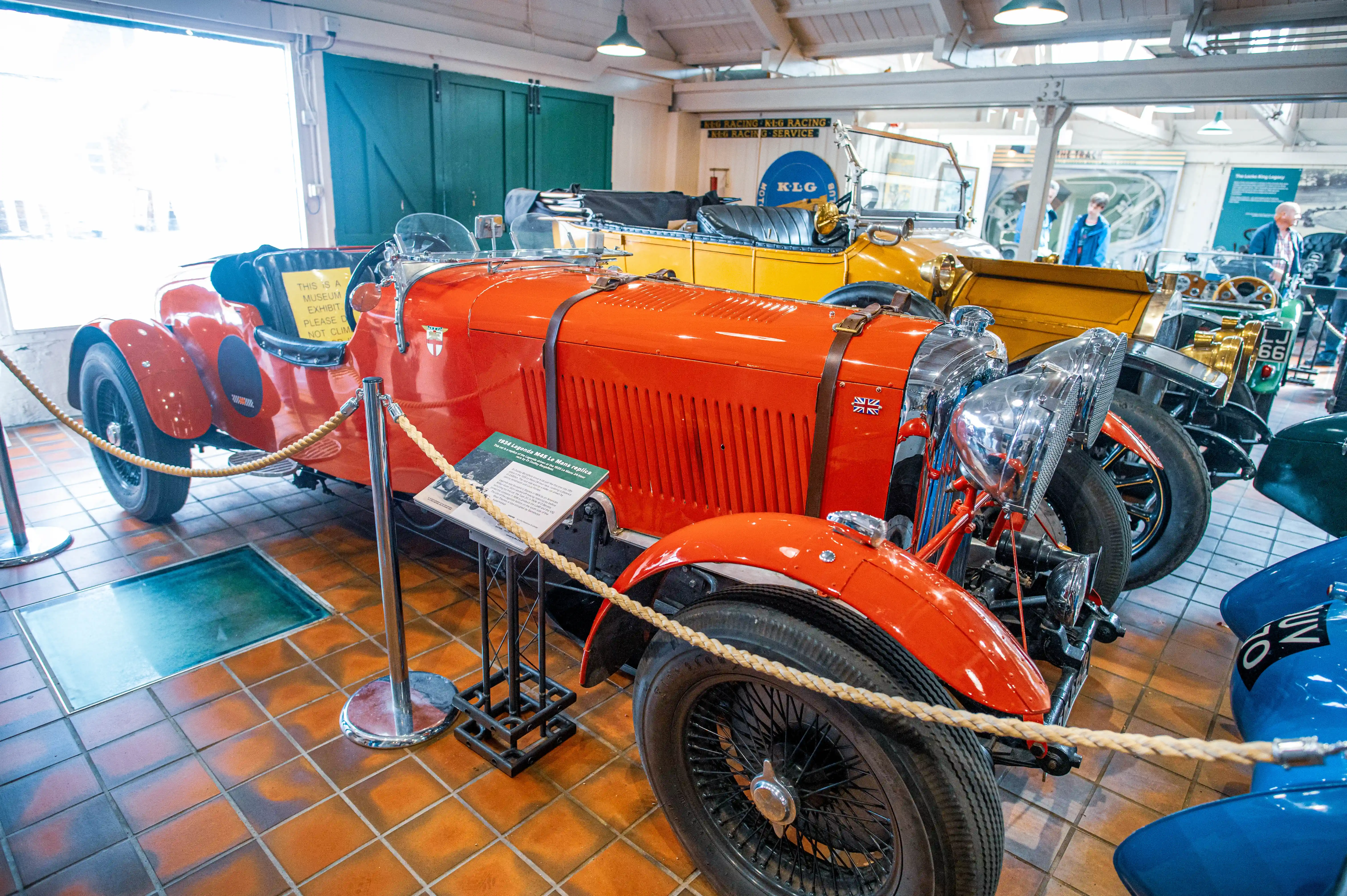 A bright orange vintage racing car is parked indoors, with a rope barrier in front of it. A sign on the driver's seat reads THIS IS A MUSEUM EXHIBIT. PLEASE DO NOT CLIMB. Other vintage cars, including a yellow convertible and a green car with the number 56 on its licence plate, are visible in the background.