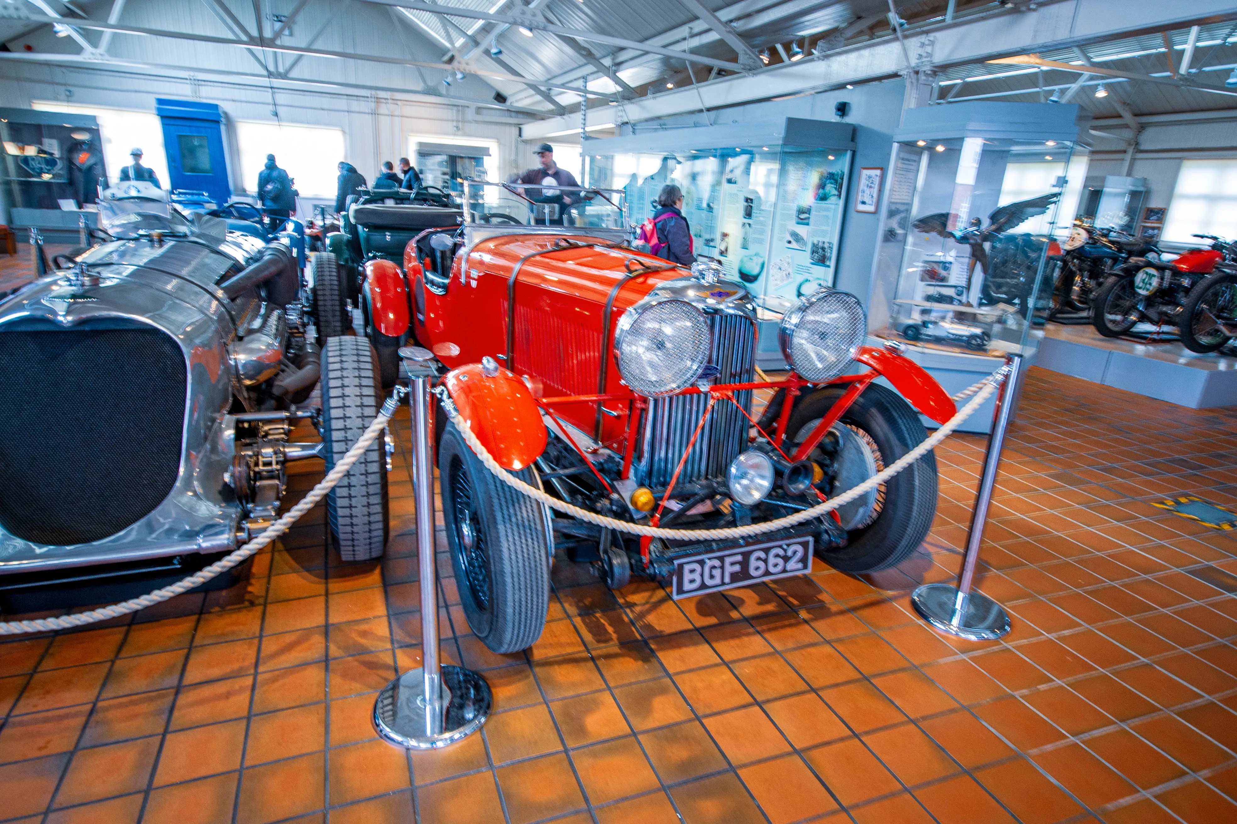 A front view of a bright red vintage open-top sports car with large round headlights, chrome grille, and visible engine components, displayed on a tiled floor in a museum. A silver vintage car is to its left, and other motorcycles and cars are visible further back.