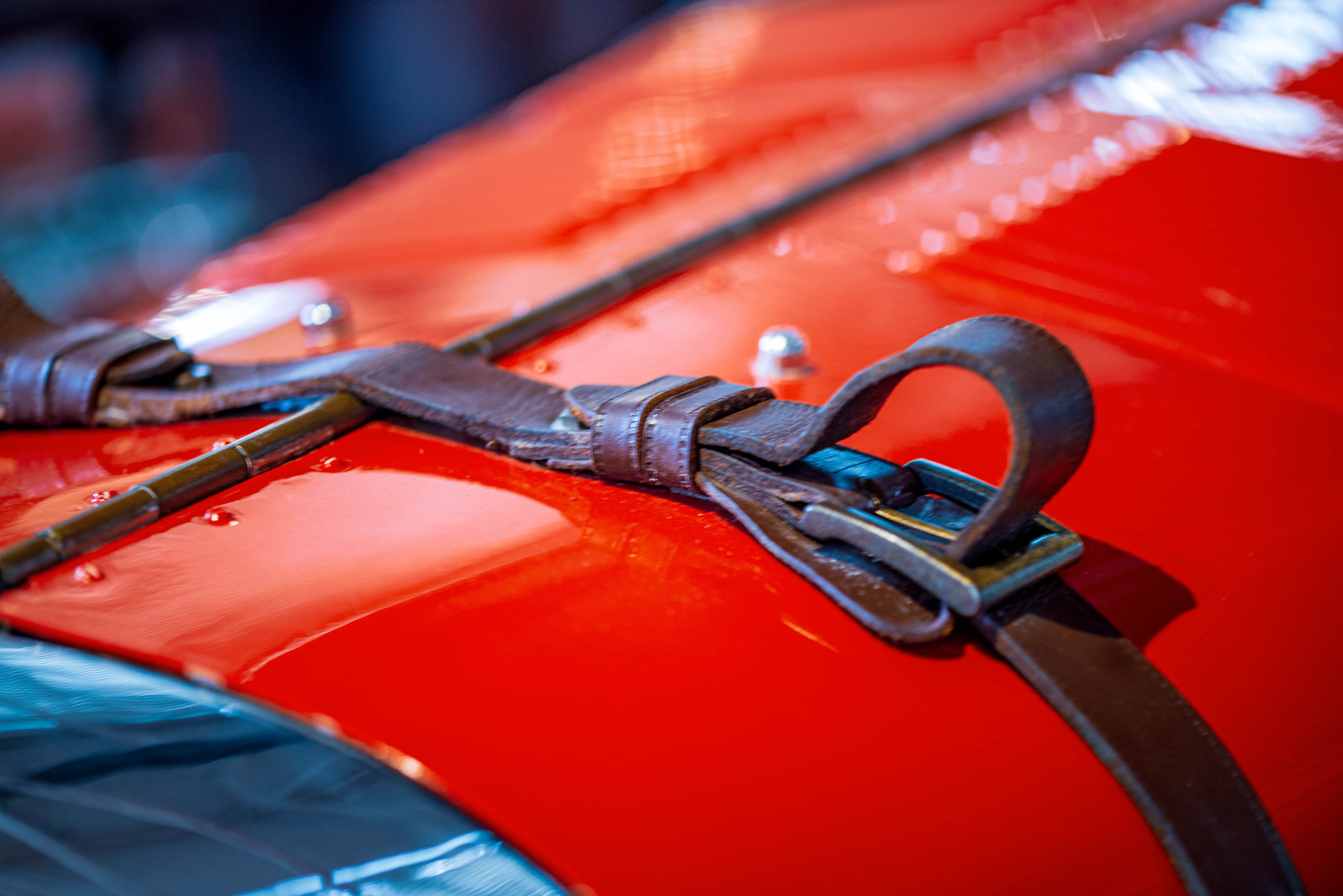 A close-up view of the hood of a vintage car, featuring brown leather straps with metal buckles securing the hood in place. The car’s vibrant red paint and curved fender are visible, highlighting the craftsmanship and attention to detail of the vehicle’s design
