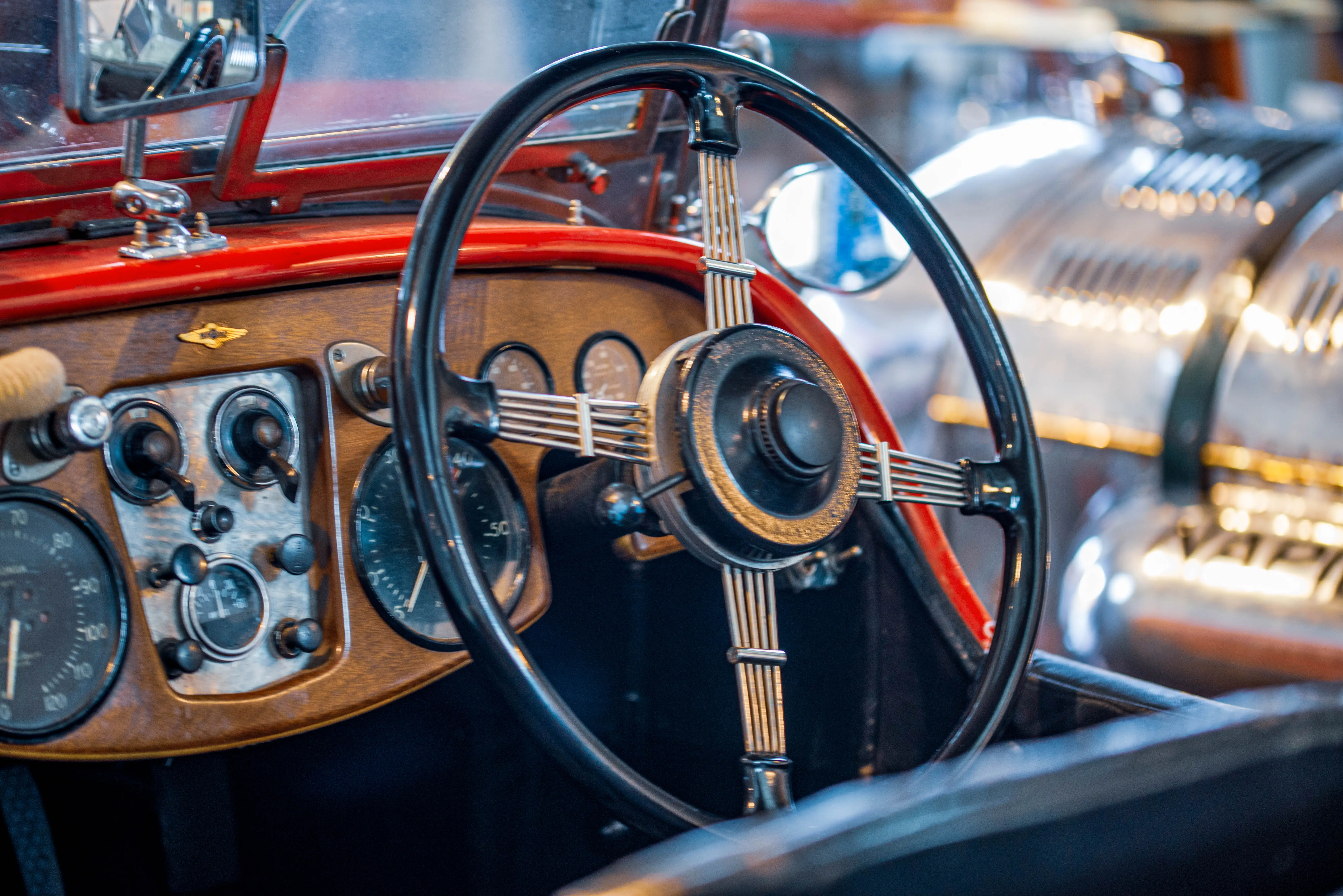 Interior view of a vintage red car, focusing on the black steering wheel with chrome spokes and the polished wooden dashboard with multiple analog gauges and switches.