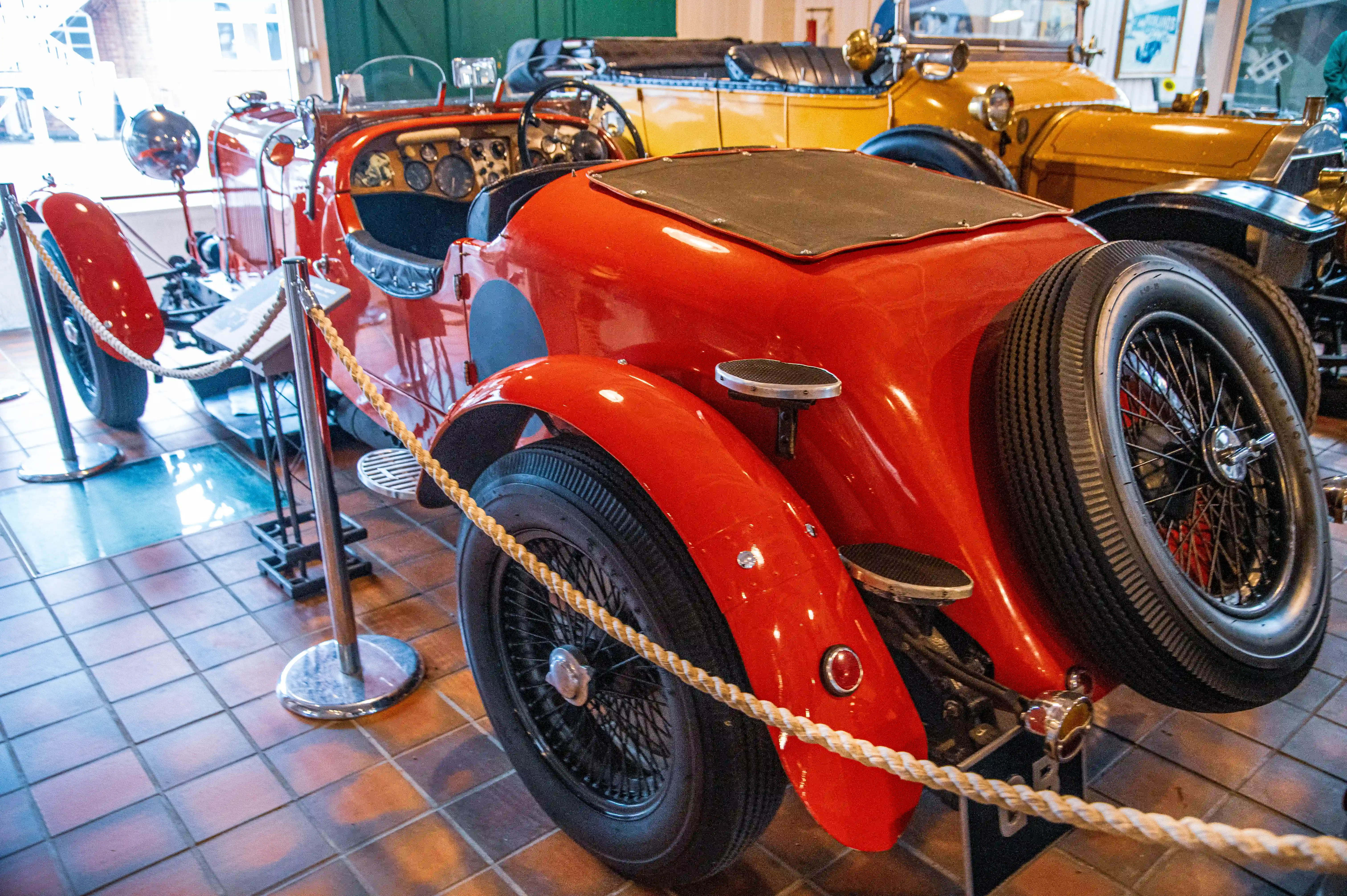 A bright red vintage sports car with a spare tyre on the back is parked indoors on a tiled floor, cordoned off with a rope. A yellow vintage car is in the background.