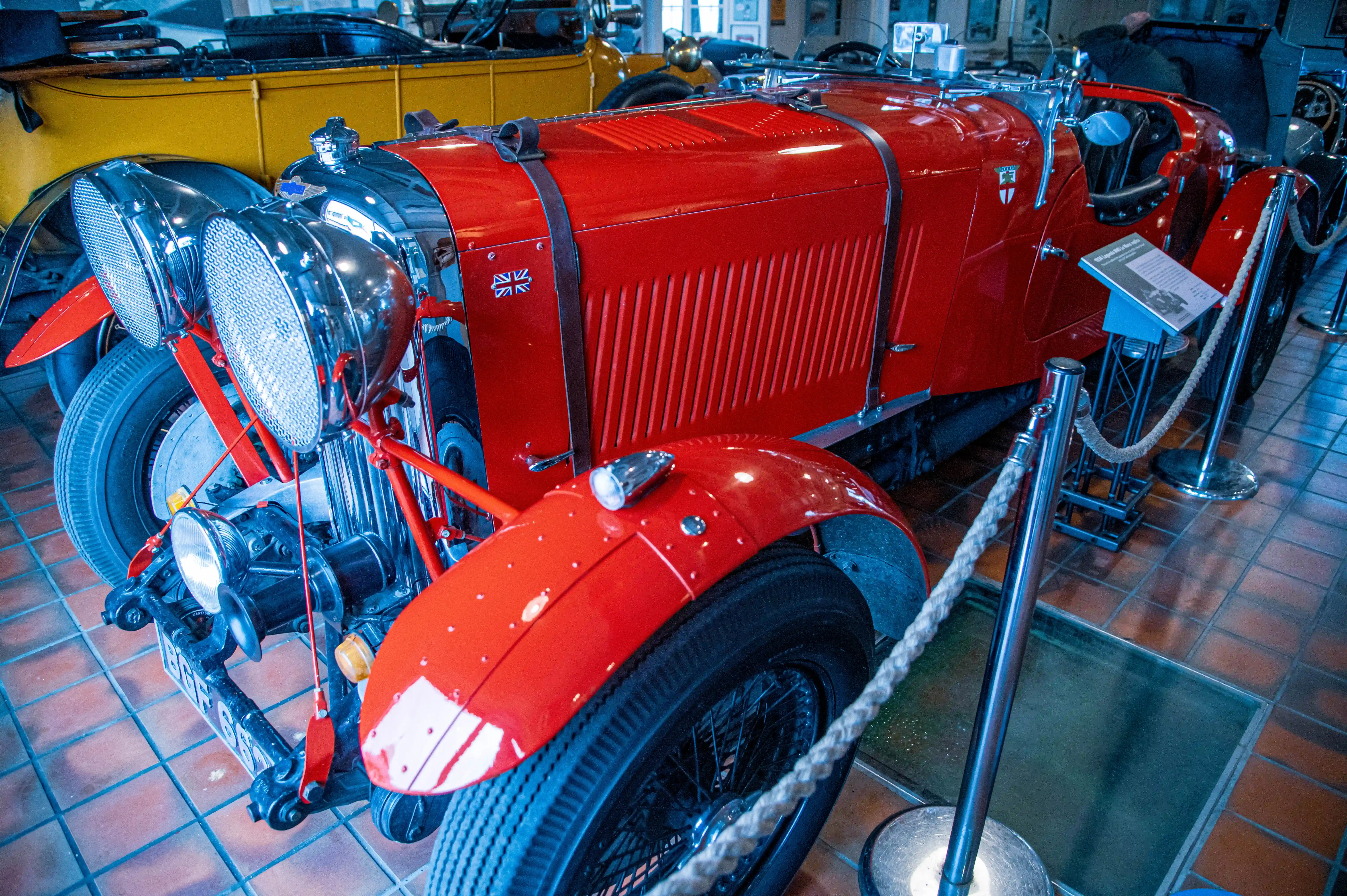 Close-up view of a bright red vintage sports car with large chrome headlights, a Union Jack flag sticker on the side panel, and wire-spoke wheels, on display in a museum.