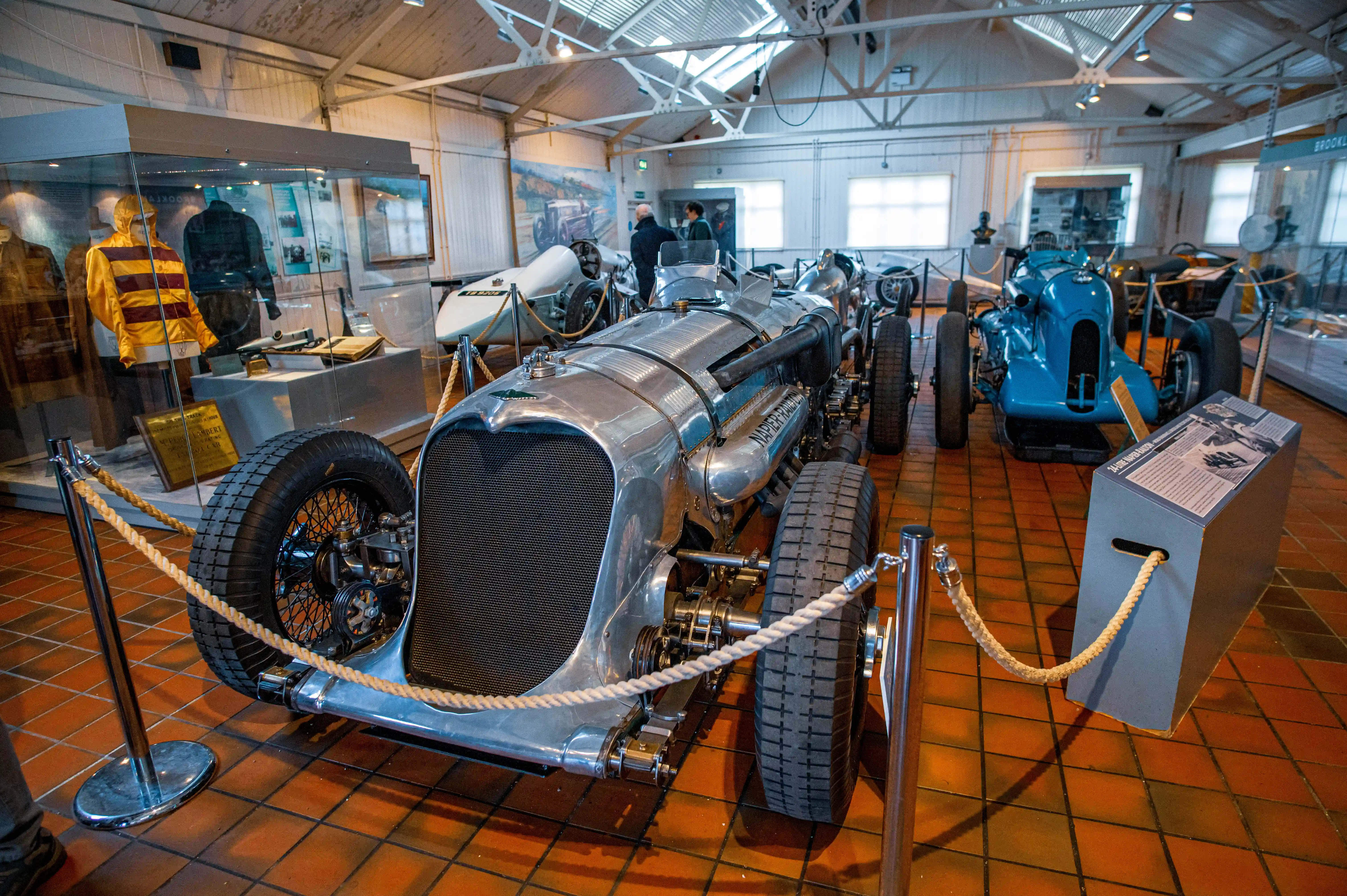 A museum interior displaying vintage racing cars, with a prominent silver Napier-Bentley in the foreground. A blue Bugatti and a white racing car are visible in the background. A display case with racing gear is to the left, and informational panels are placed near the cars.