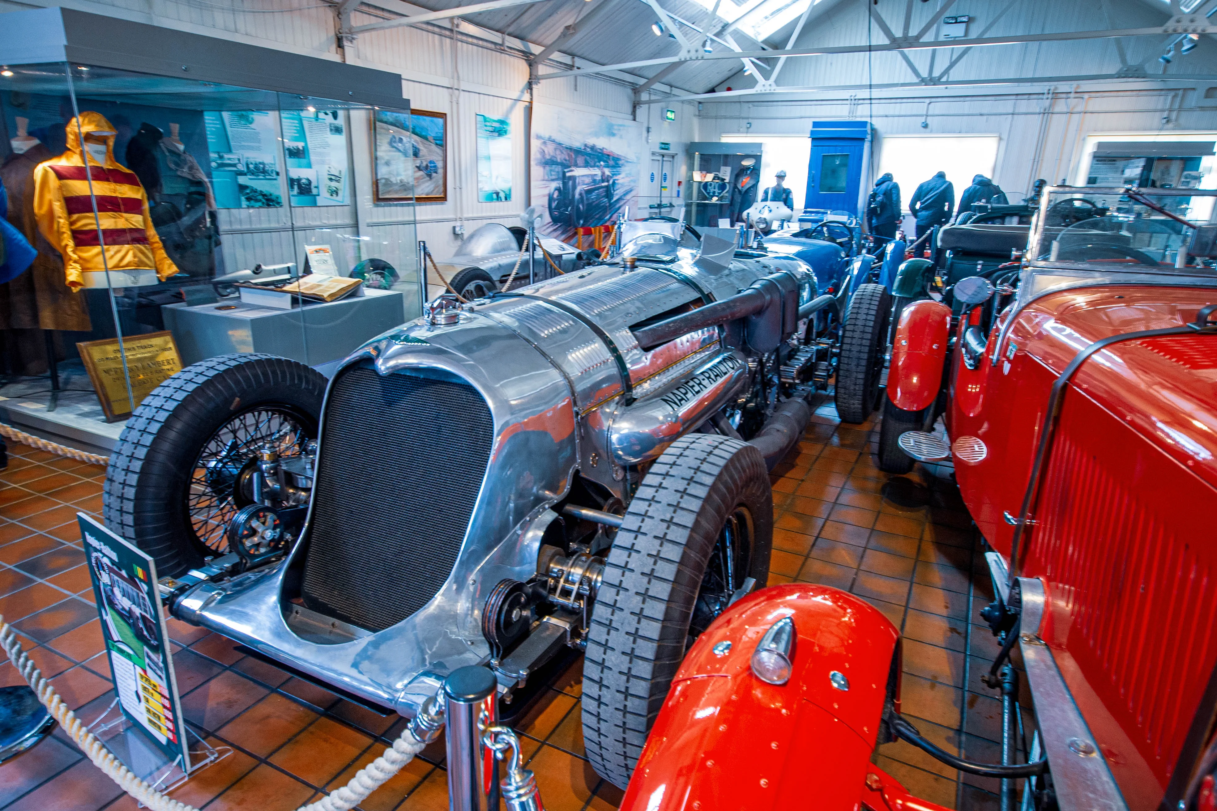 A museum exhibition hall featuring several vintage racing cars. The centrepiece is a gleaming silver Napier-Railton car with exposed engine components and a wire-spoked wheel. To the right, part of a bright red classic sports car is visible. In the background, other cars and display cases containing historical items and mannequins wearing racing gear can be seen.