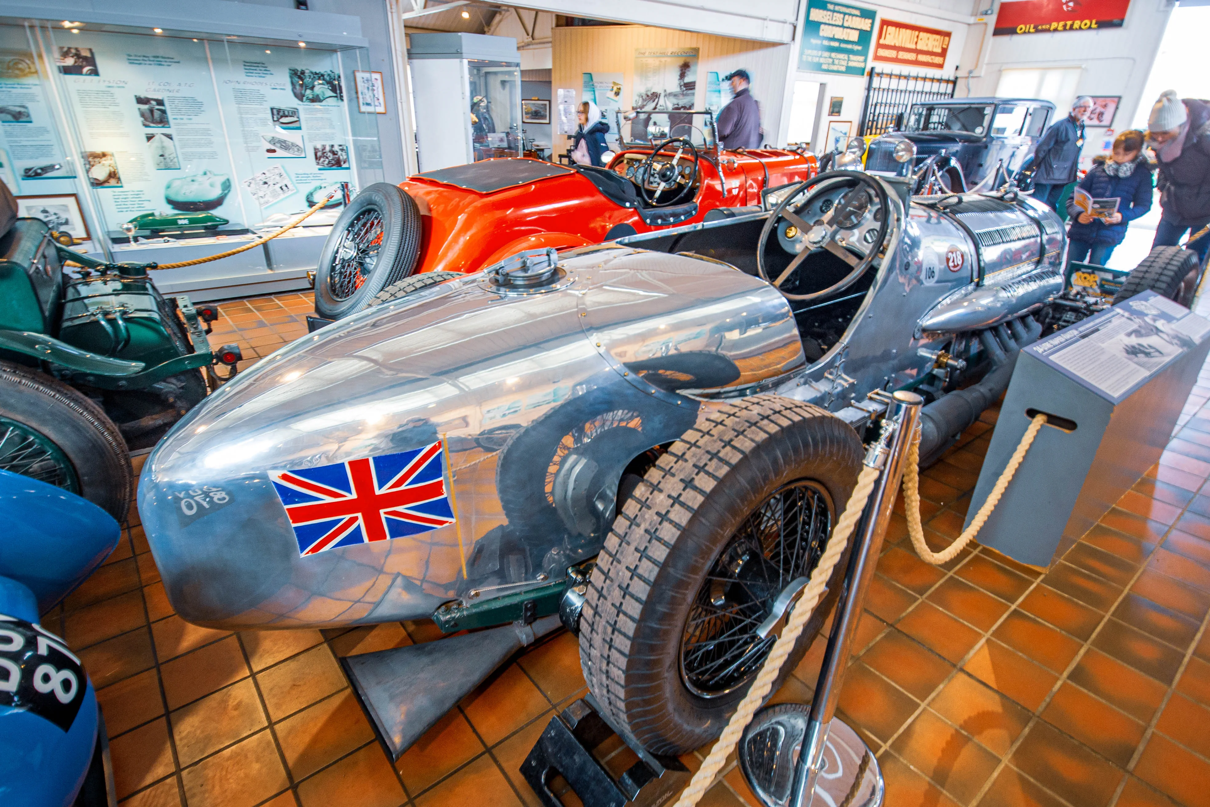A close-up view of a polished silver vintage racing car with a Union Jack decal on its side, parked in a museum alongside other classic automobiles.