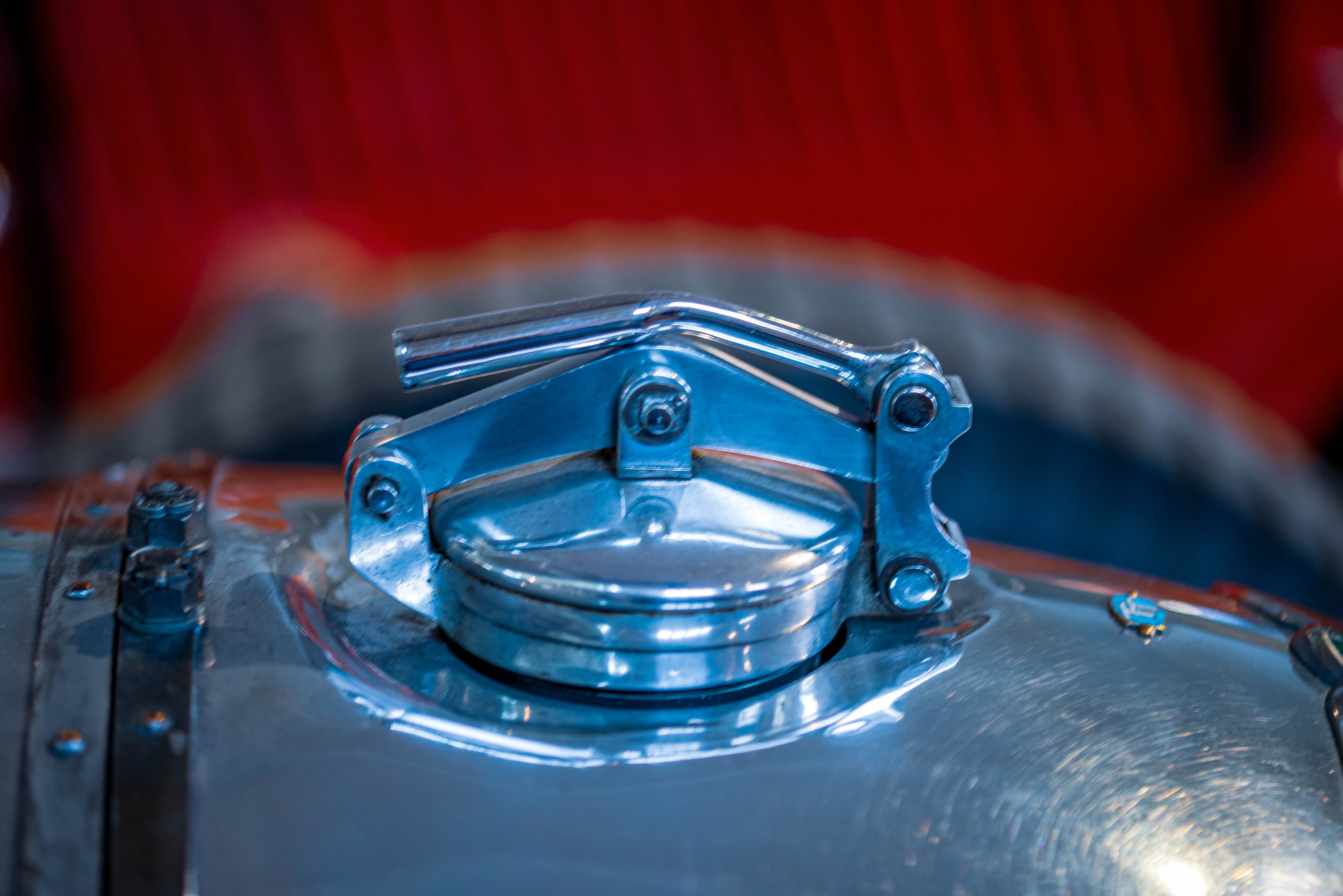 Detailed view of a shiny chrome fuel cap and its hinged latch on a vintage car, with a blurred red background.