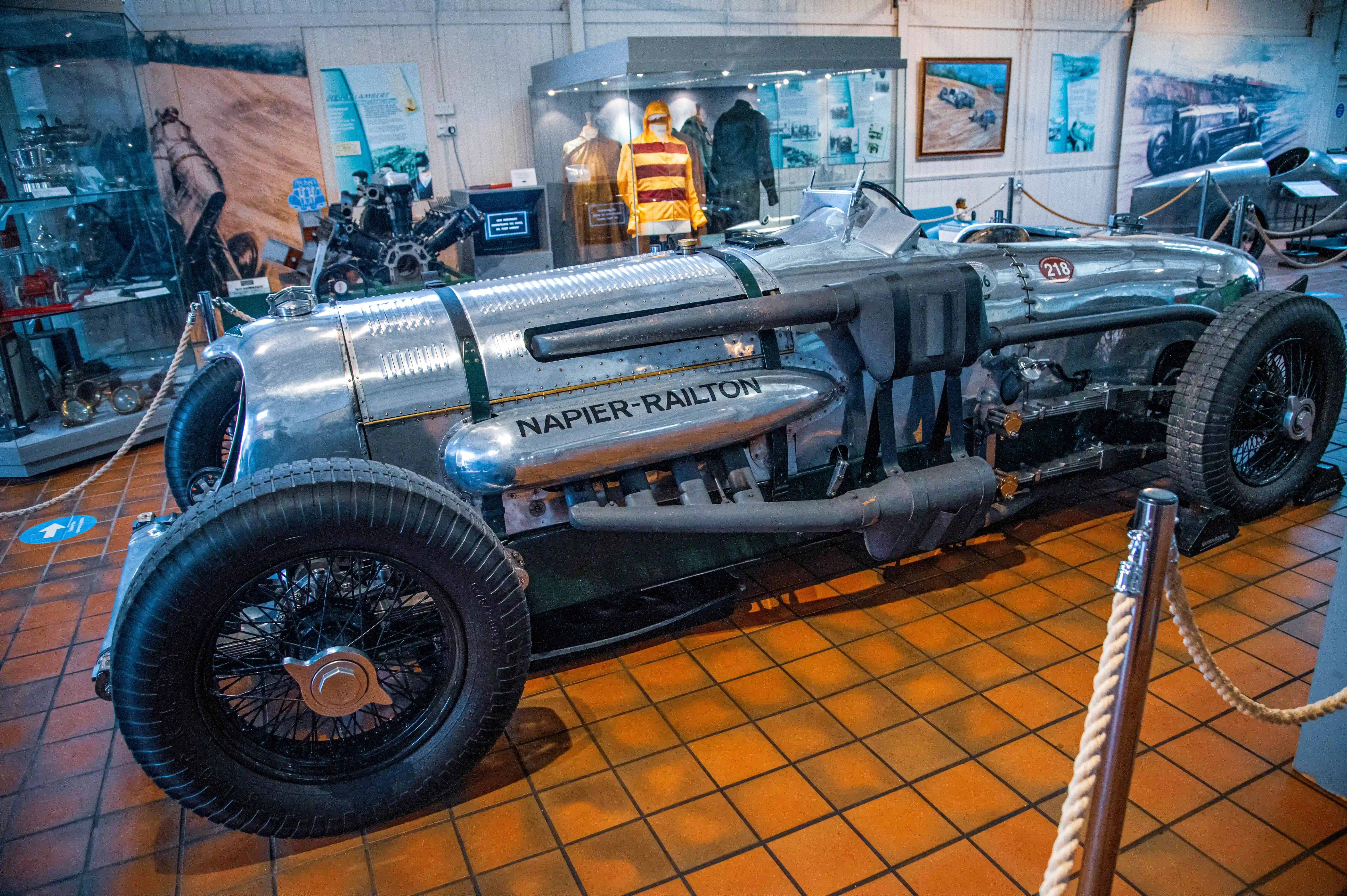 A vintage Napier-Railton racing car with the name NAPIER-RAILTON emblazoned on its silver fuselage, displayed on a tiled floor in a museum.