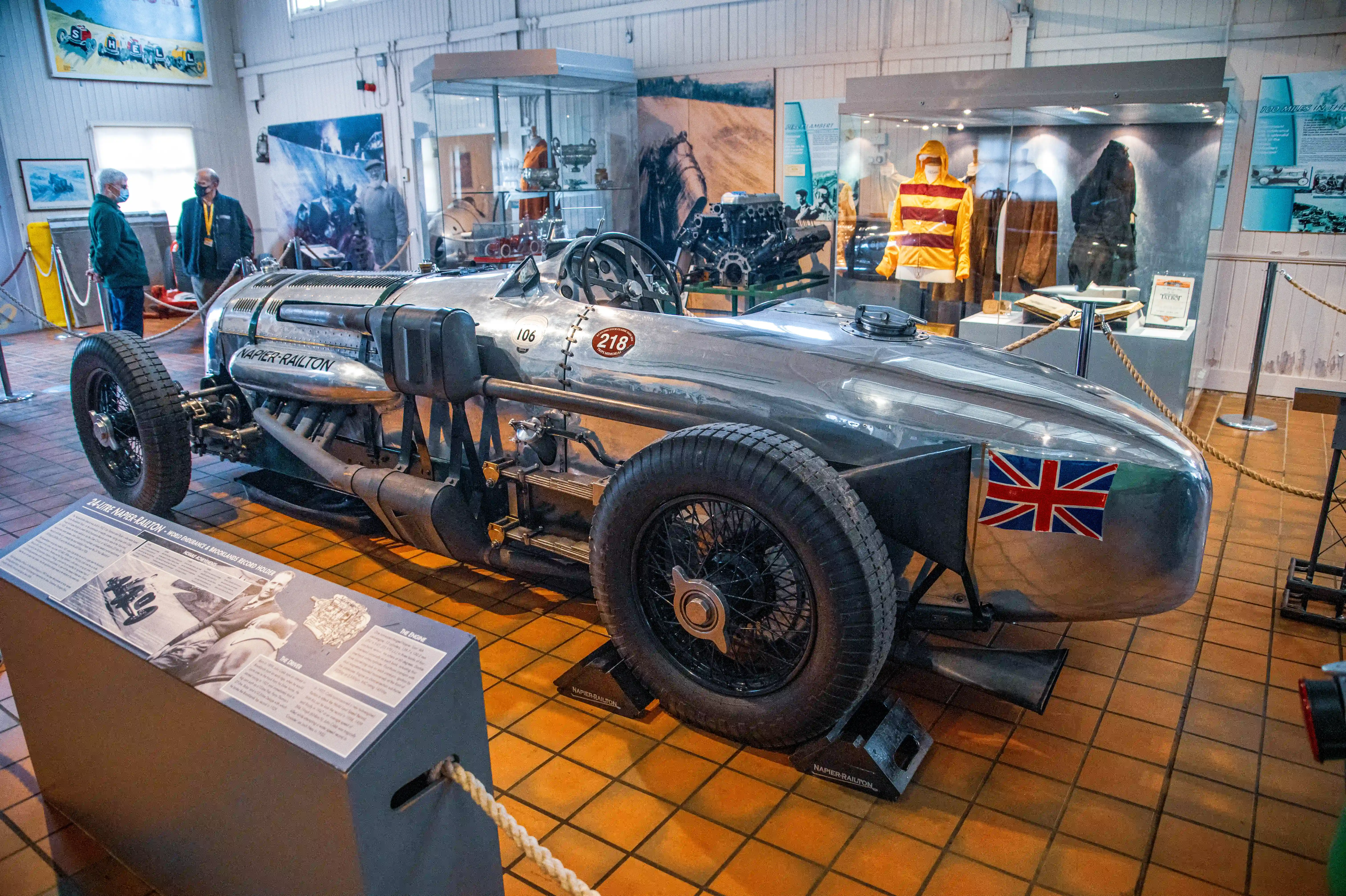 A close-up, low-angle view of the silver Napier-Railton land speed record car in a museum. The car has wire-spoked wheels, a prominent engine, and a Union Jack flag on its rear. Two people in masks are visible in the background, looking at the car. A display board in the foreground provides information about the vehicle.