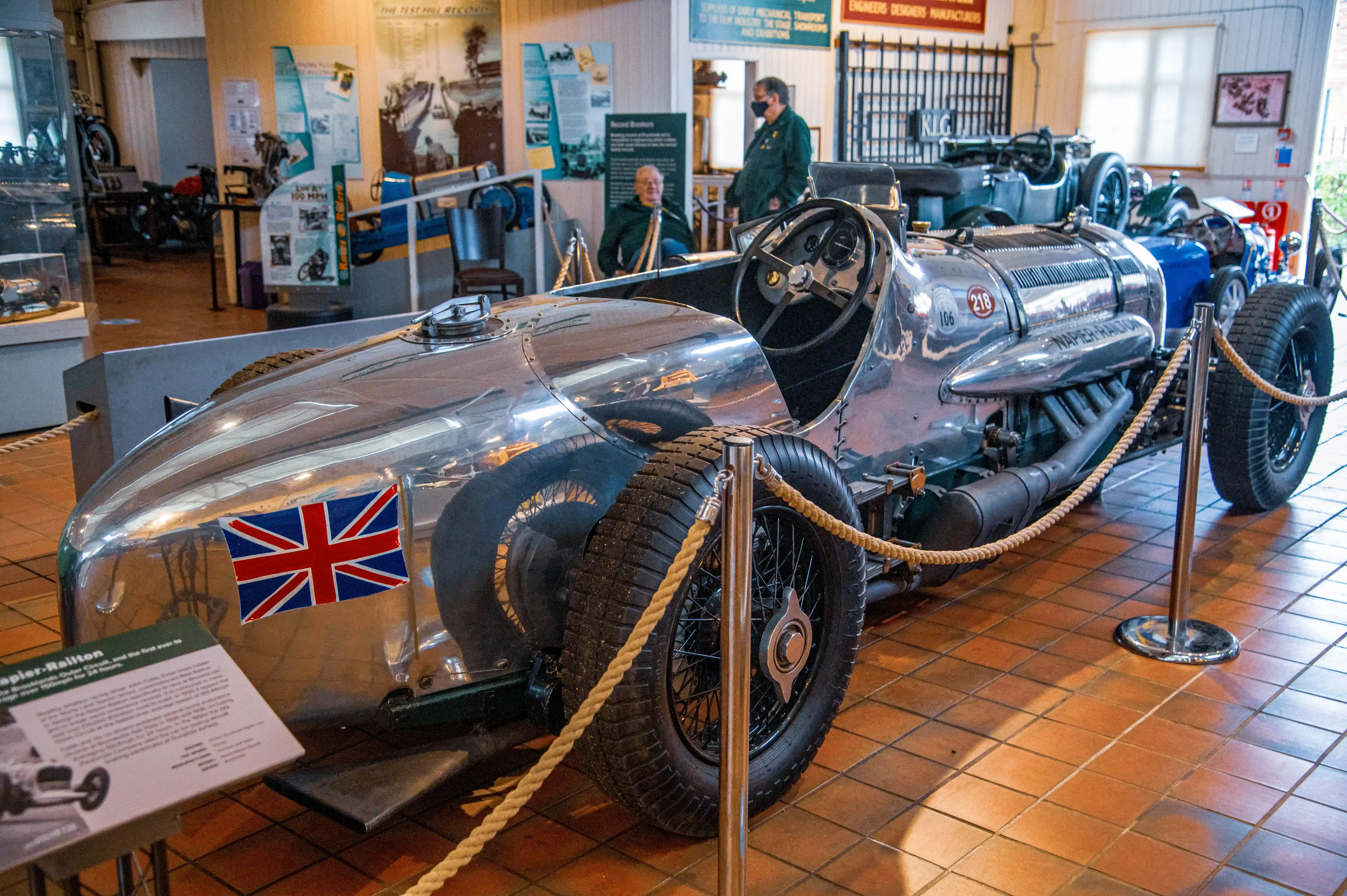 A polished silver Napier-Railton racing car with a Union Jack flag on its nose is on display in a museum. The car has a large engine, spoked wheels, and is cordoned off with rope barriers. Two men are visible in the background, one in a green jacket and the other in a dark top.