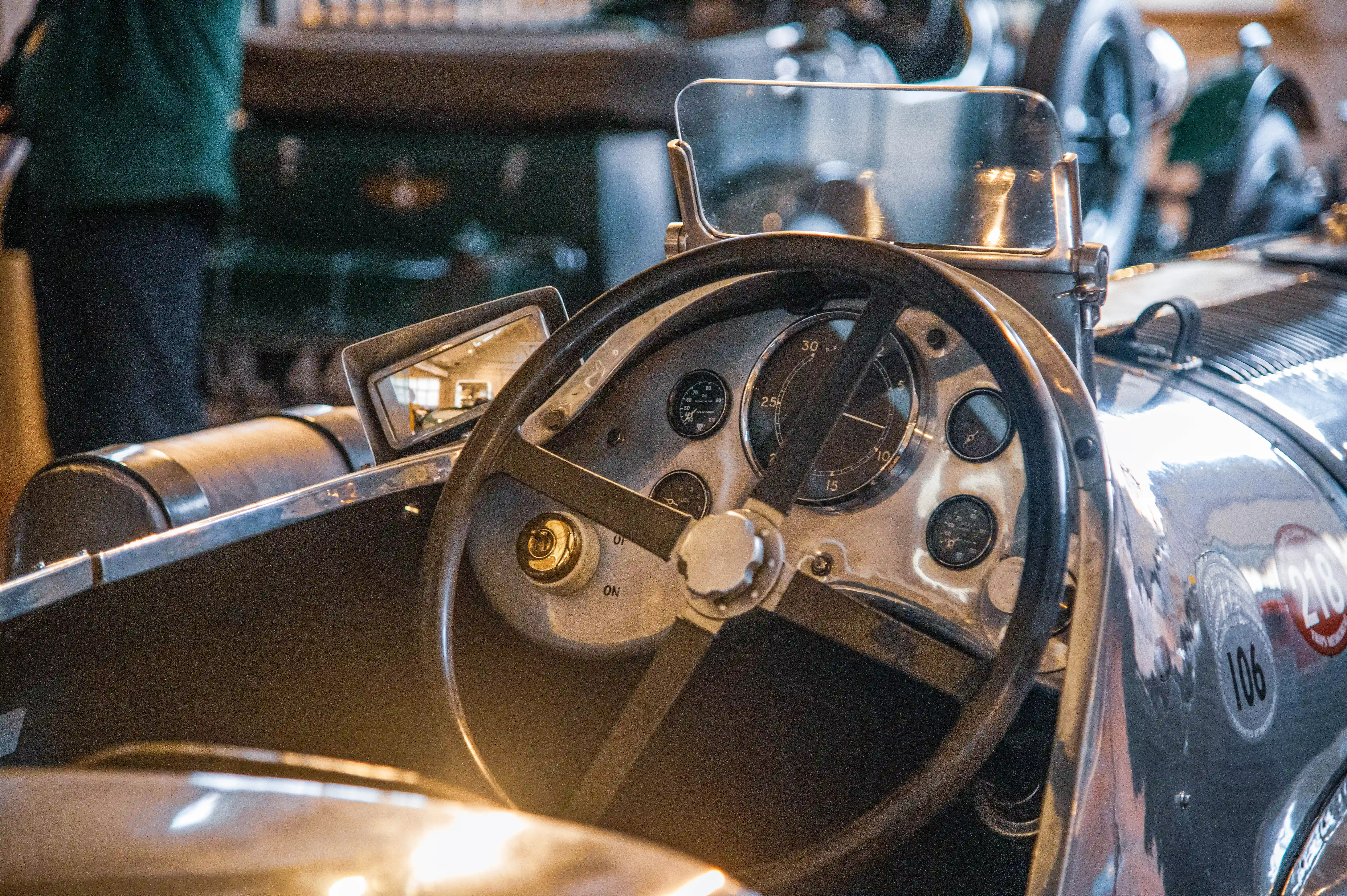 Close-up of a vintage racing car's interior, focusing on the steering wheel, dashboard with analogue gauges, and a polished metal body.