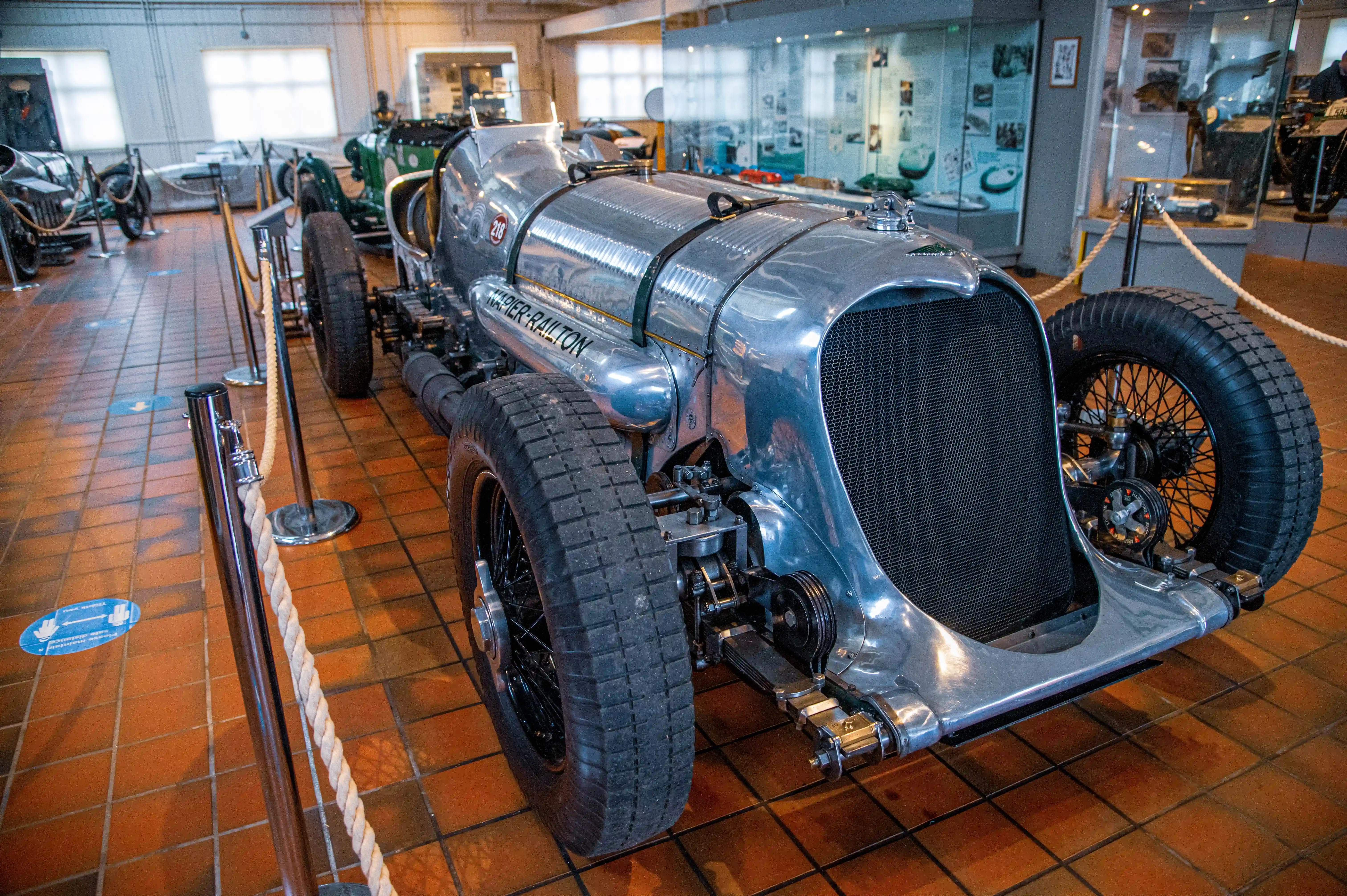 Front quarter view of the silver Napier-Railton land speed record car in a museum setting, showcasing its historic design, including its large grille, spoked wheels, and the 