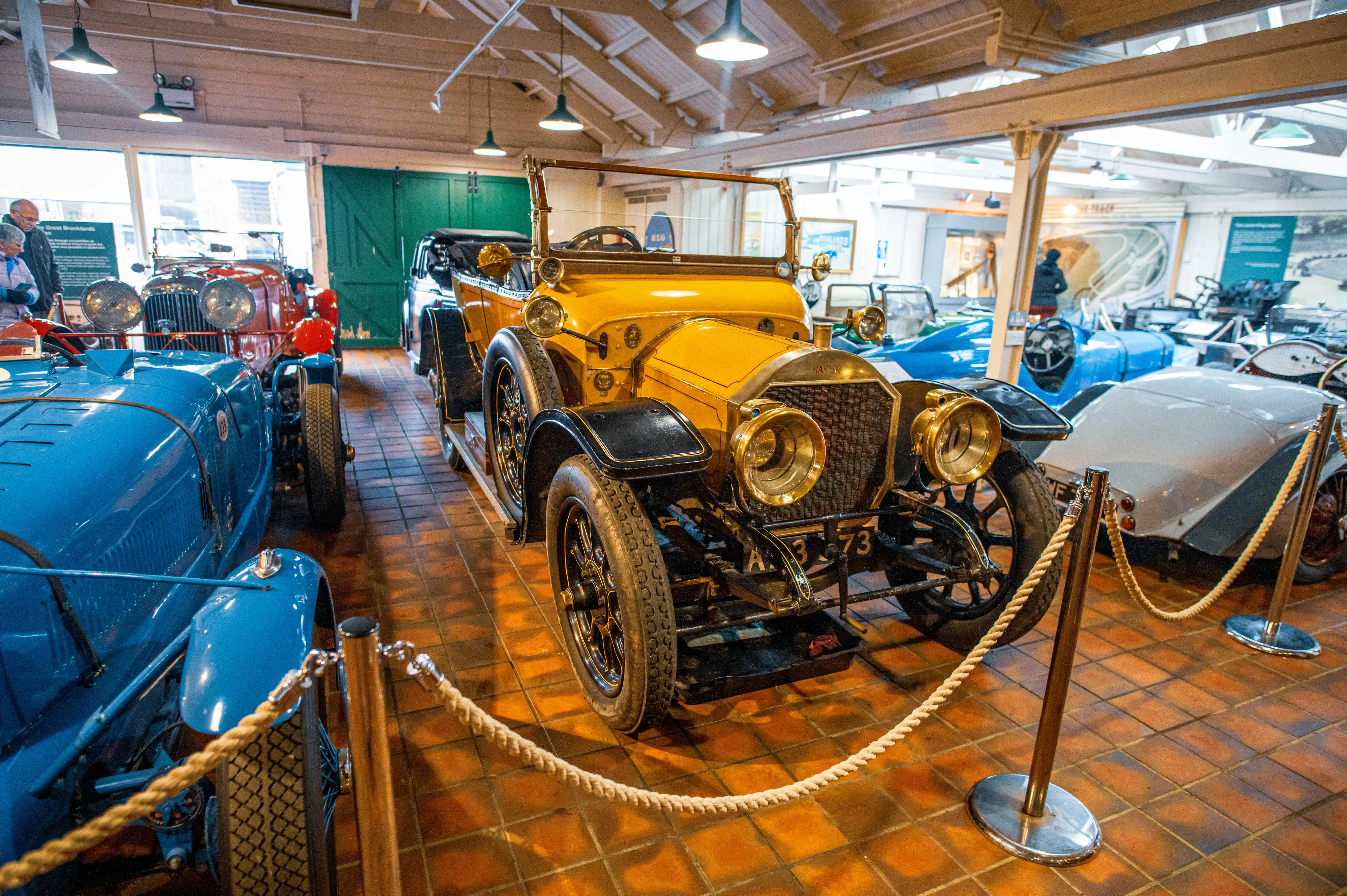 A collection of vintage cars on display in a museum. The foreground features a bright yellow Napier with brass accents and wire-spoke wheels. Behind it, a blue open-top car and a light grey vintage car are partially visible. The cars are arranged on a tiled floor, with rope barriers in the foreground.