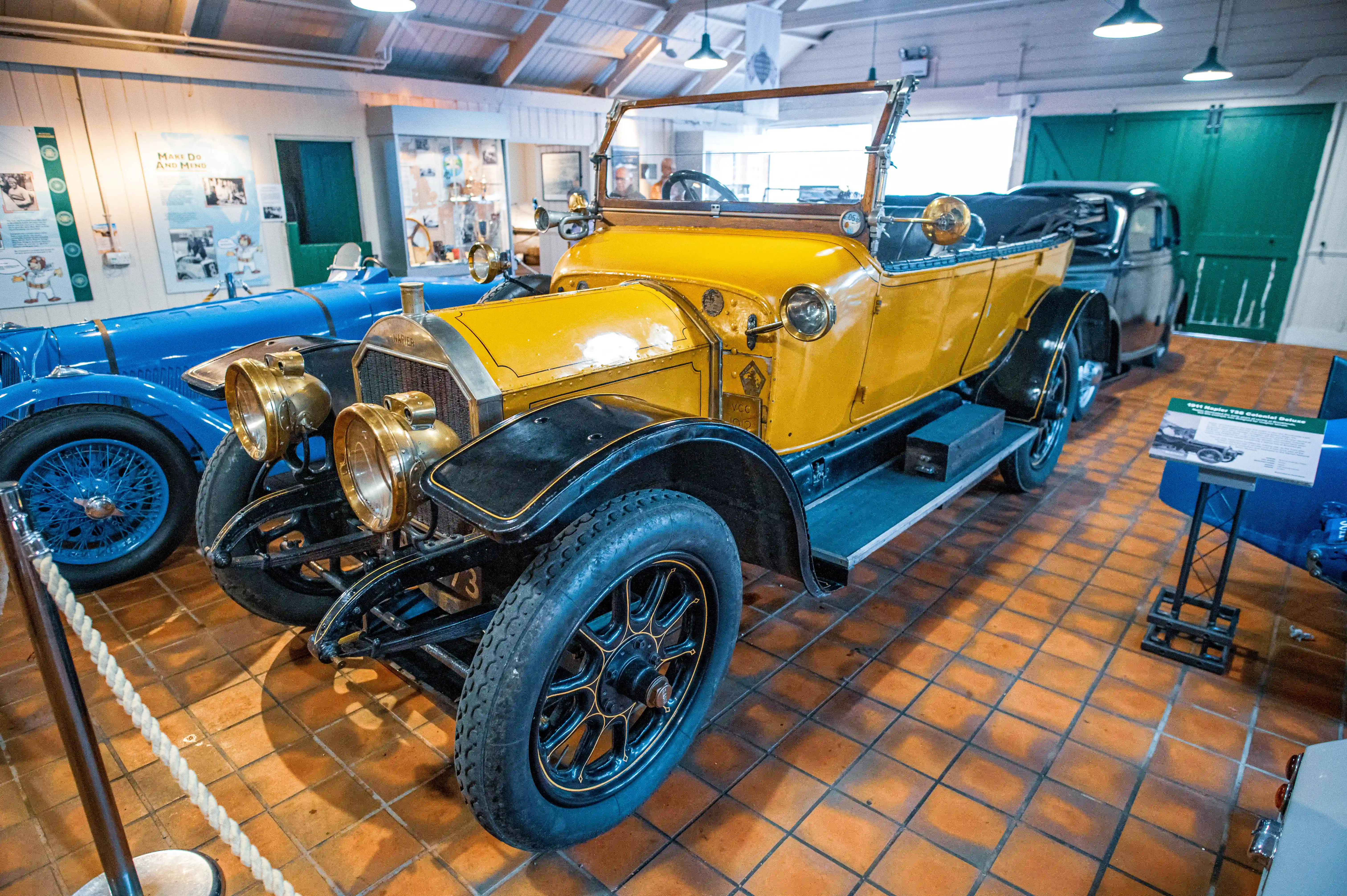 A vibrant yellow 1911 Napier Colonial Deluxe convertible car with brass headlamps is displayed prominently in a museum, with a blue vintage racing car beside it. The cars are on a tiled floor.