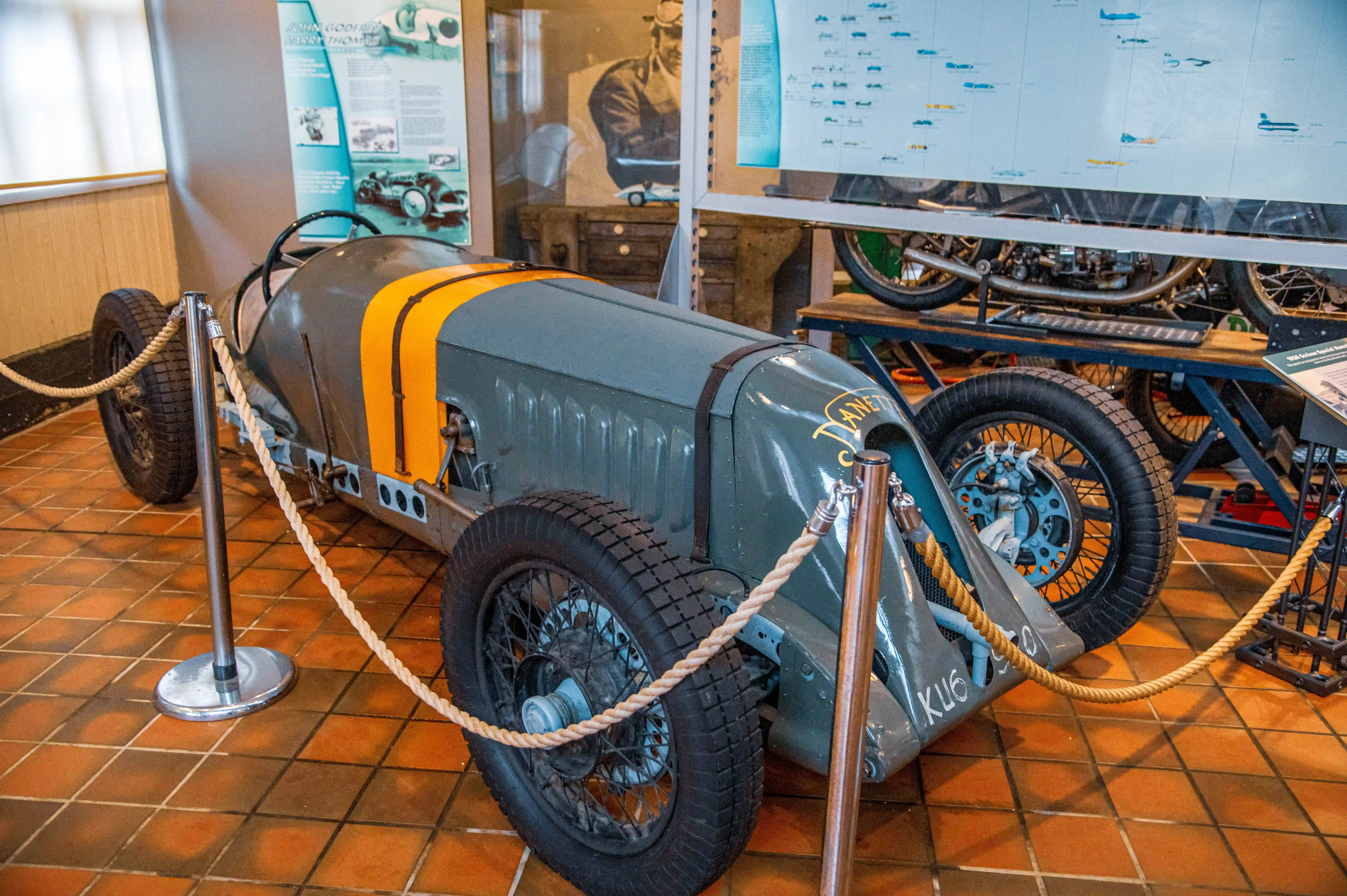 A grey vintage racing car with orange stripes and wire wheels is showcased in a museum setting, with informational displays and another vehicle in the background.