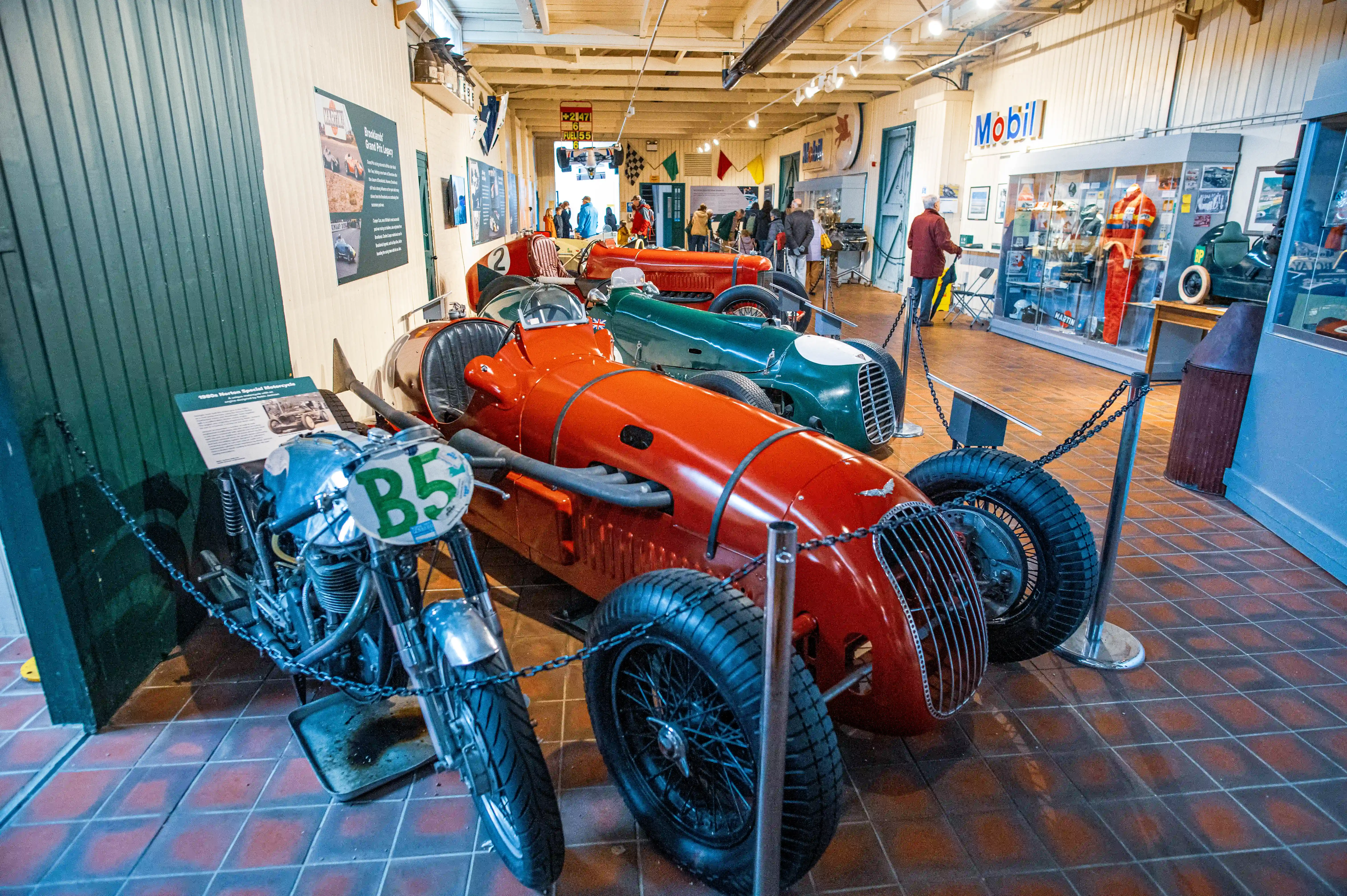  A bright red vintage racing car with a prominent grille and wire-spoked wheels sits on a tiled floor in a museum. A silver motorcycle is behind it, and a green racing car is to the right. A visitor in a blue jacket is in the background.
