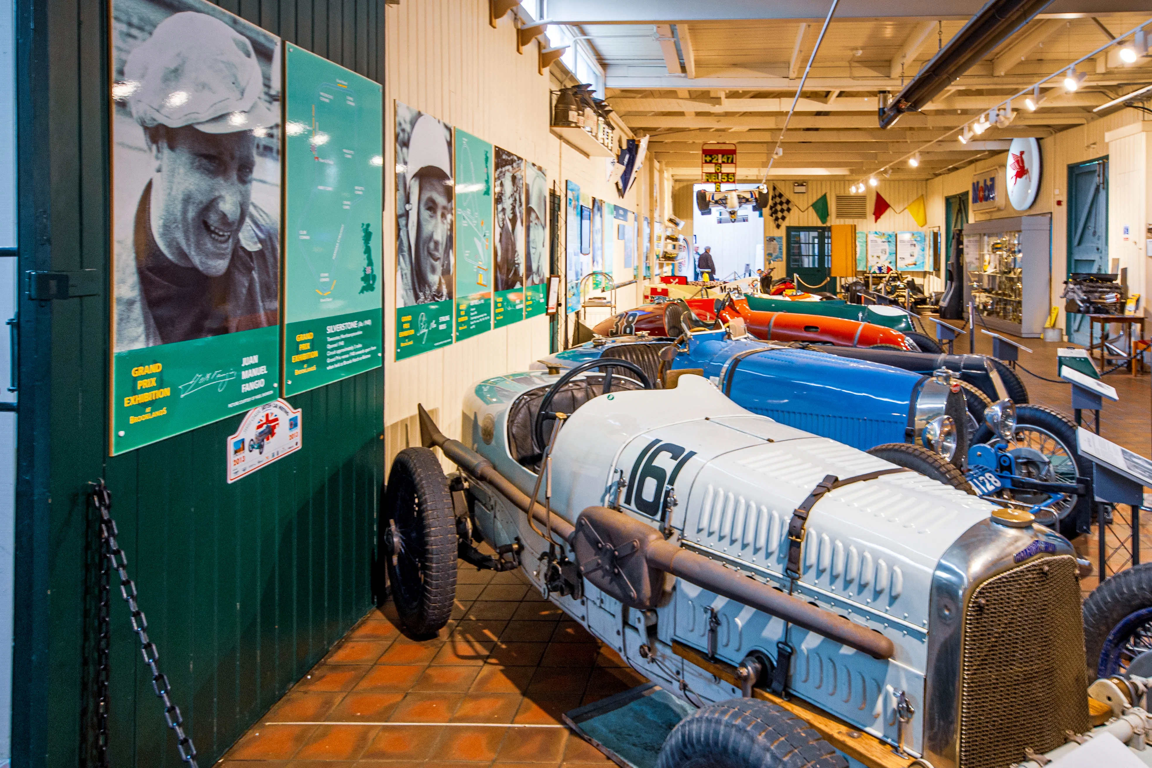 A museum exhibition hall displaying several vintage racing cars, with a light blue Bugatti prominently in the foreground. Large posters of a smiling Juan Manuel Fangio and racing circuits adorn a green wall to the left.