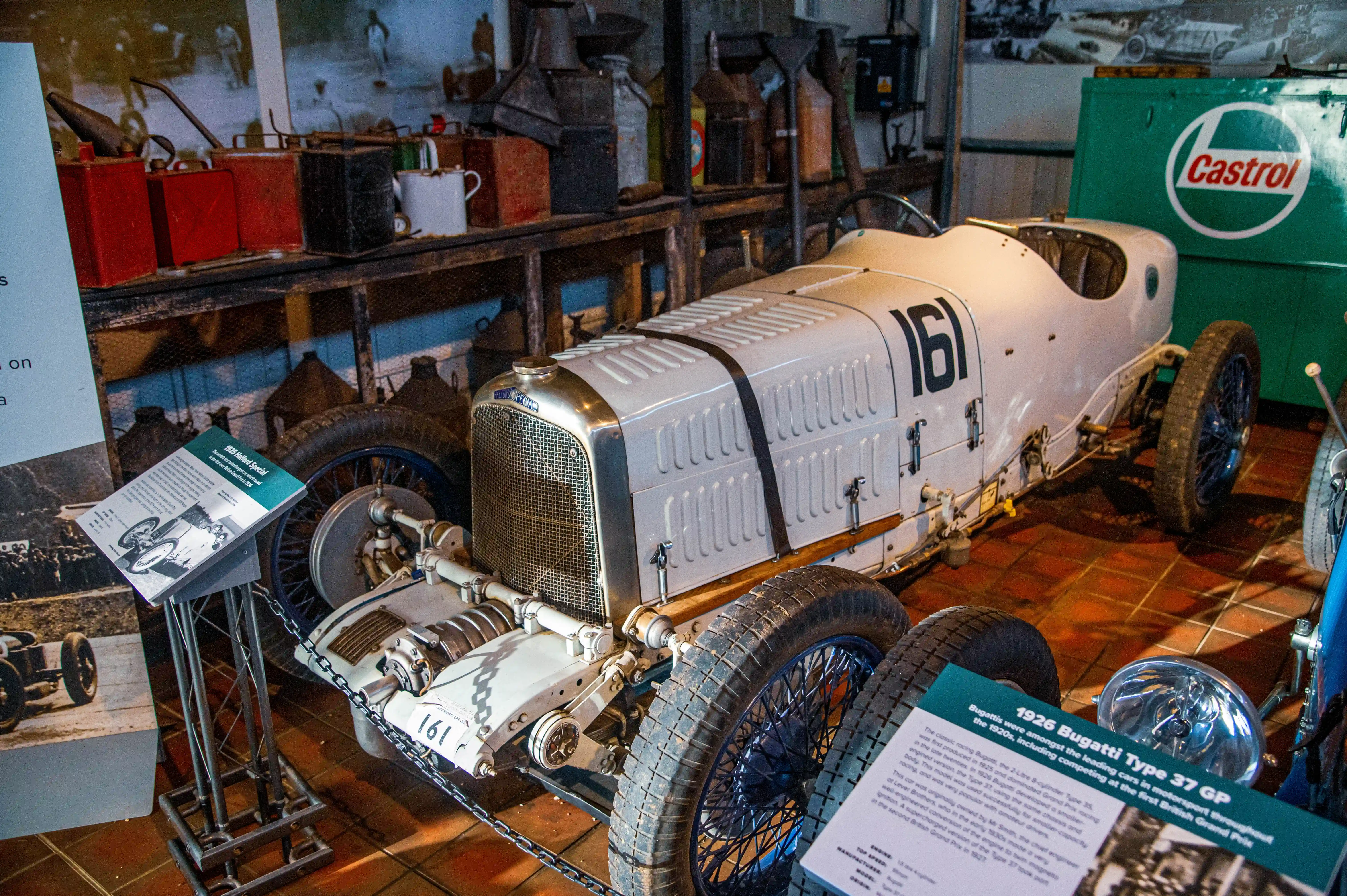 A vintage 1926 Bugatti Type 37 GP racing car, painted white with the number 161 on its side, is parked indoors. The car has wire-spoke wheels and a prominent grille. Various vintage motoring items, such as red and black petrol cans and oil containers, are visible on shelves behind the car, along with a large green Castrol sign.