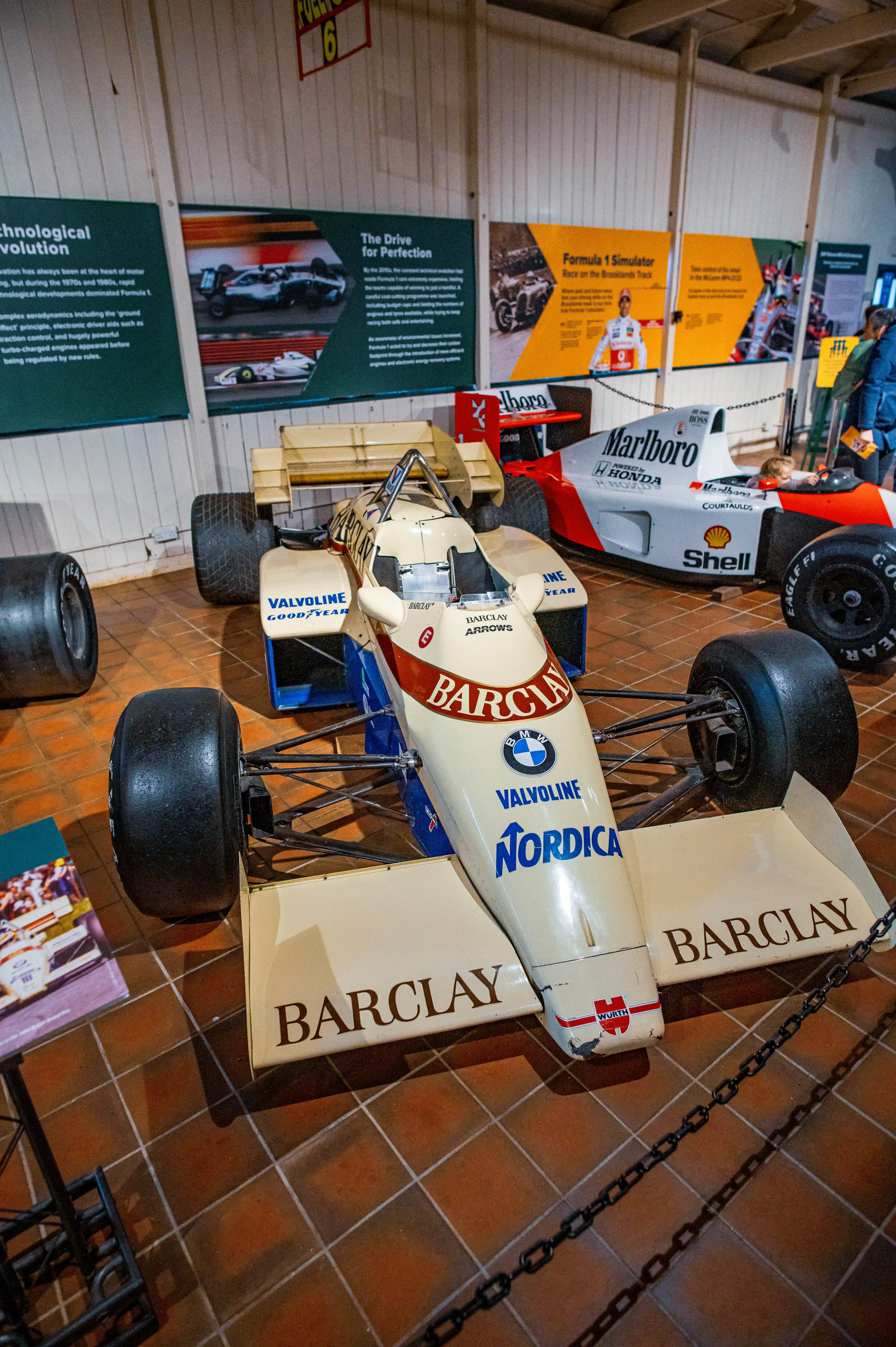 Two Formula 1 racing cars are displayed indoors on a tiled floor. The car in the foreground is an open-wheel design with a cream, blue, and red paint scheme, featuring logos such as 'Barclay', 'Valvoline', and 'Nordica'. A second car, predominantly white and red with 'Marlboro' branding, is visible behind it. Informational displays are mounted on the wall in the background.
