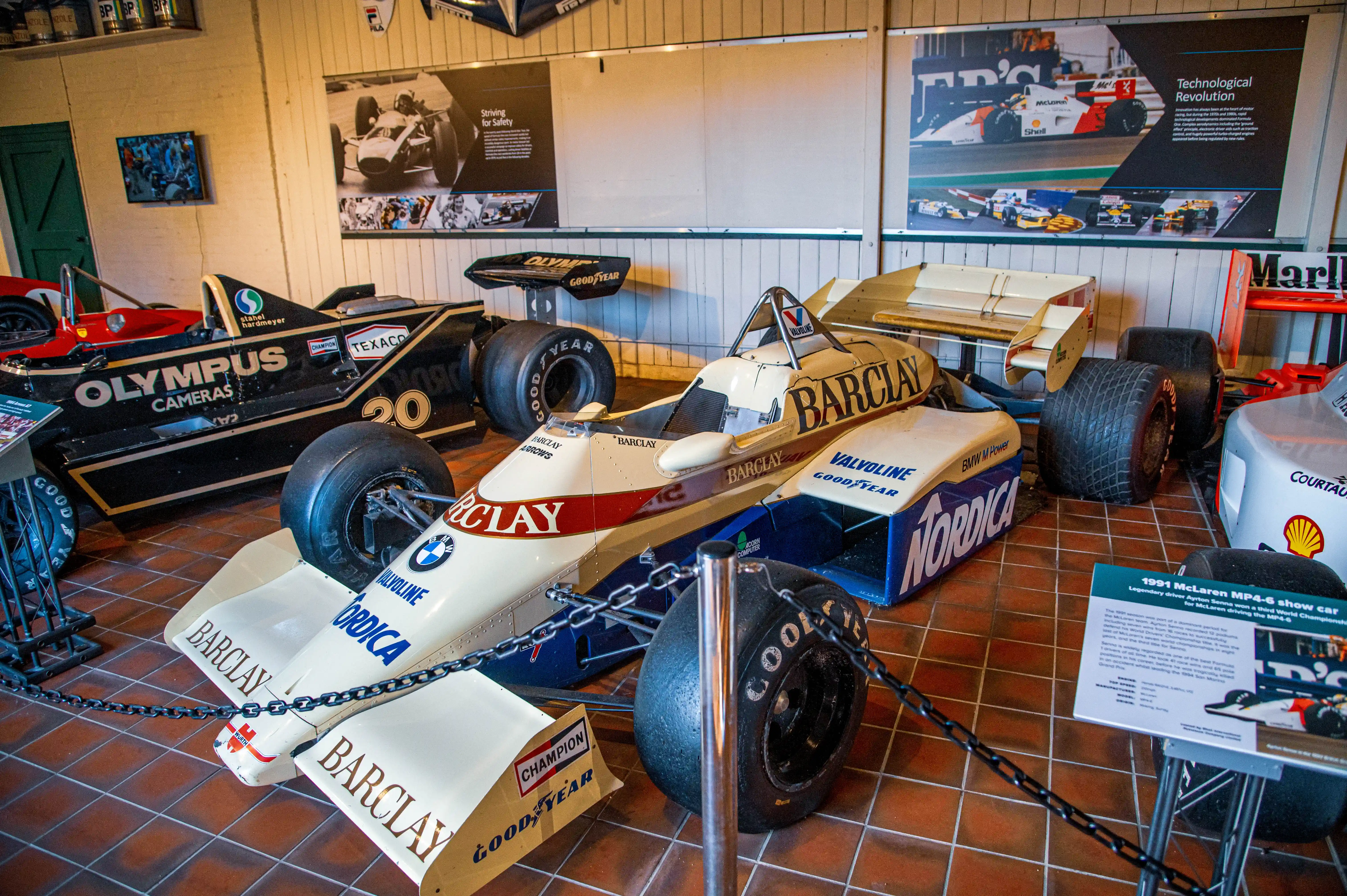 A museum display of several vintage Formula 1 racing cars, with a focus on the front section of a yellow and blue car with BARCLAY and NORDICA branding, and a portion of a black and gold car labelled OLYMPUS CAMERAS.