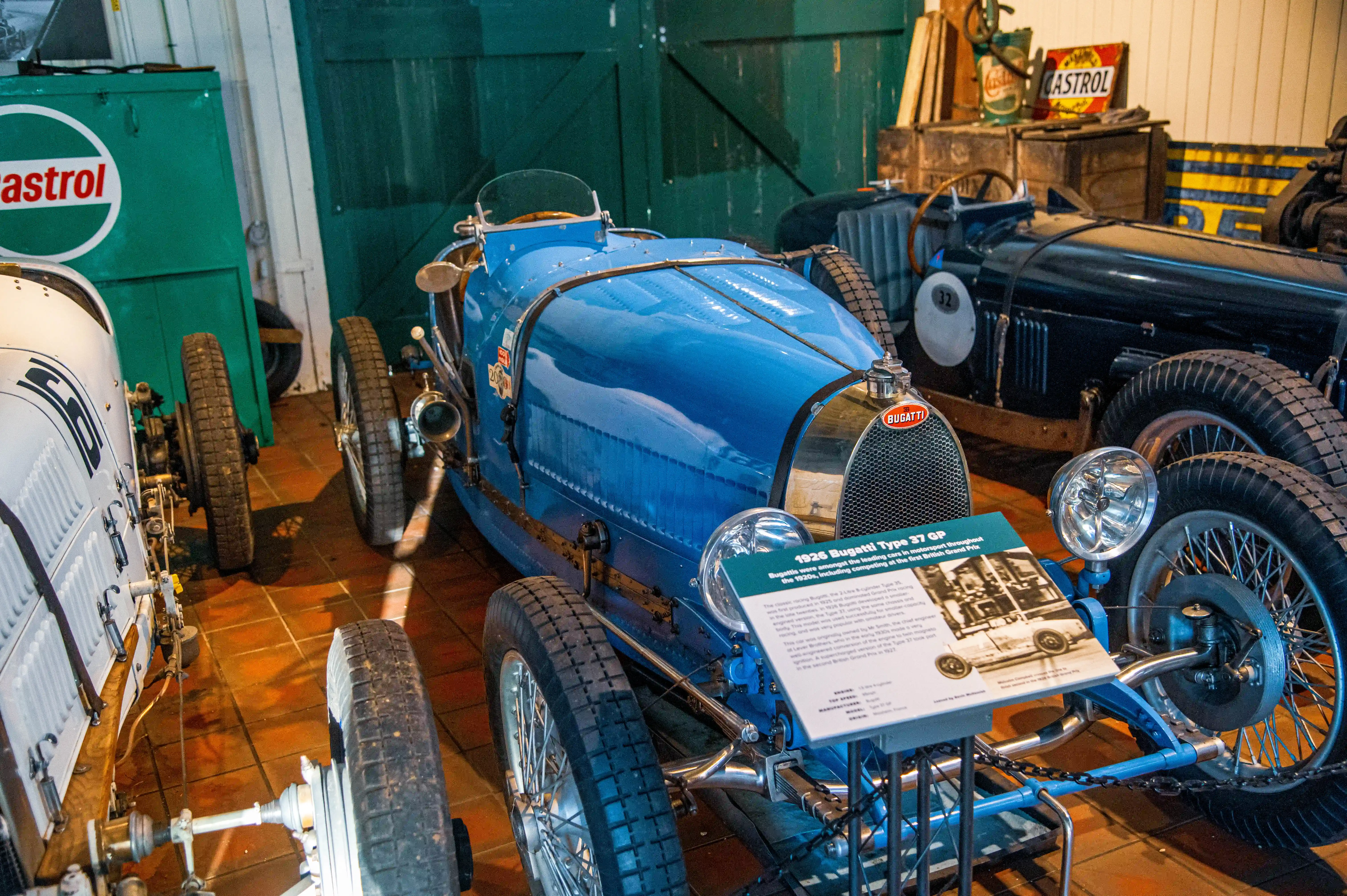 Close-up of a bright blue 1926 Bugatti Type 37 GP racing car with a sign detailing its history. Other vintage cars and garage equipment are visible in the background.