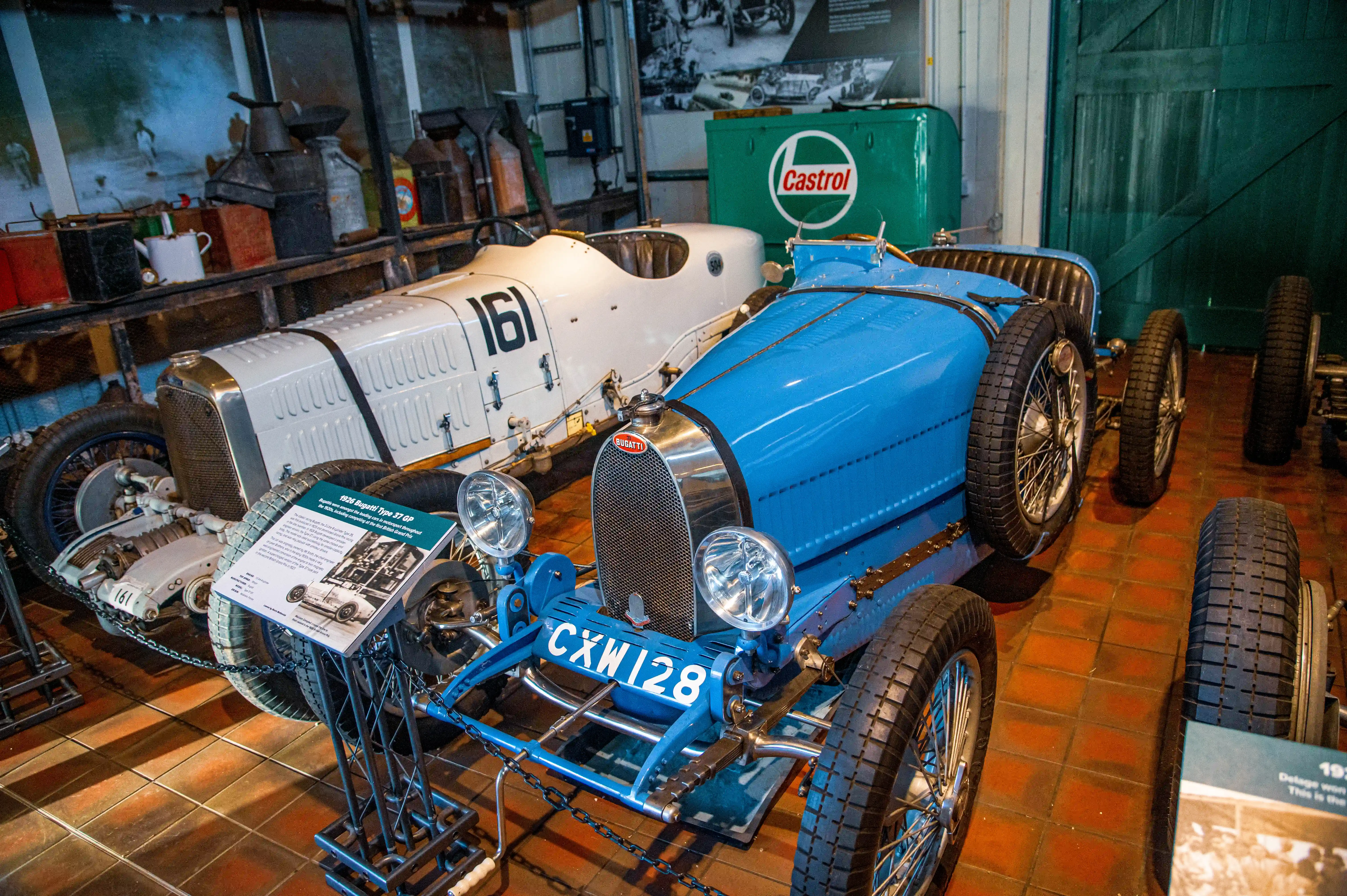 Two vintage Bugatti racing cars, one blue and one white with the number 161, are parked indoors. A green Castrol drum is in the background.