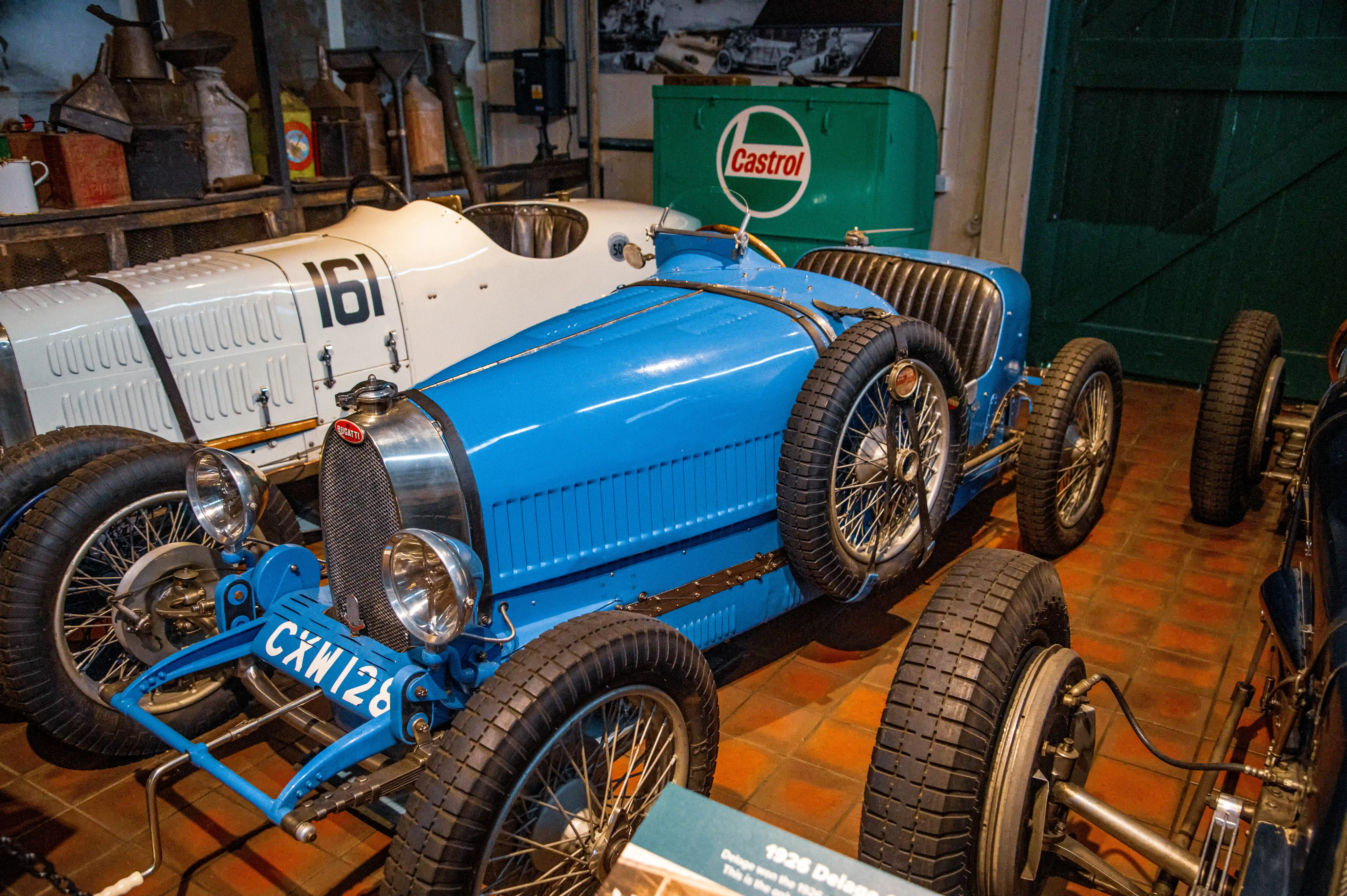 Two vintage Bugatti racing cars, one blue and one white with the number 161, are parked indoors. A green Castrol drum is in the background.