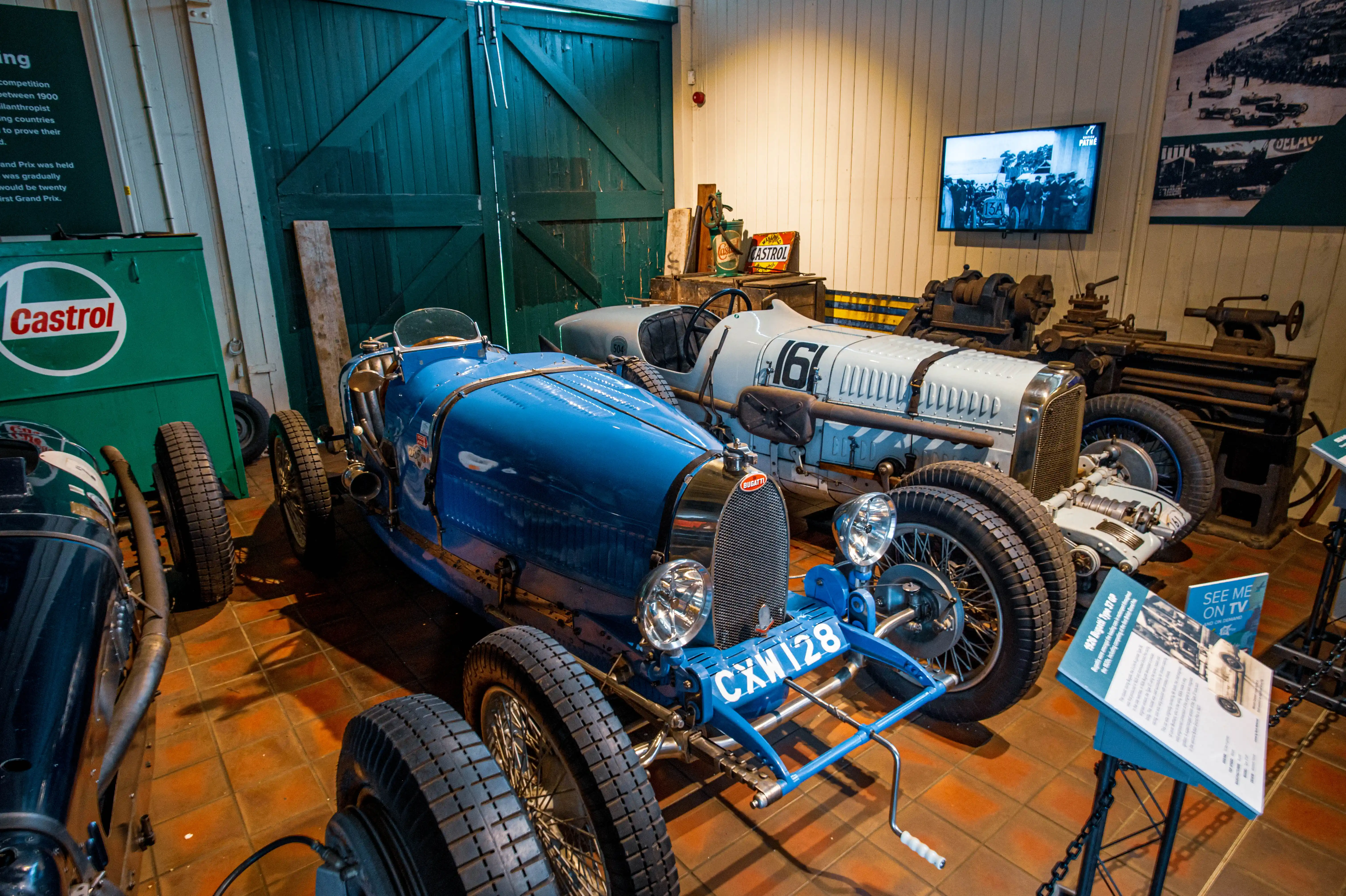 Two vintage Bugatti race cars are parked side-by-side in a museum. The car in the foreground is bright blue with a chrome grille and CXW 128 registration plate. The car behind it is light grey with the number '61' on its side. Other exhibits include old machinery, a Castrol oil can, and a screen showing black and white racing footage.