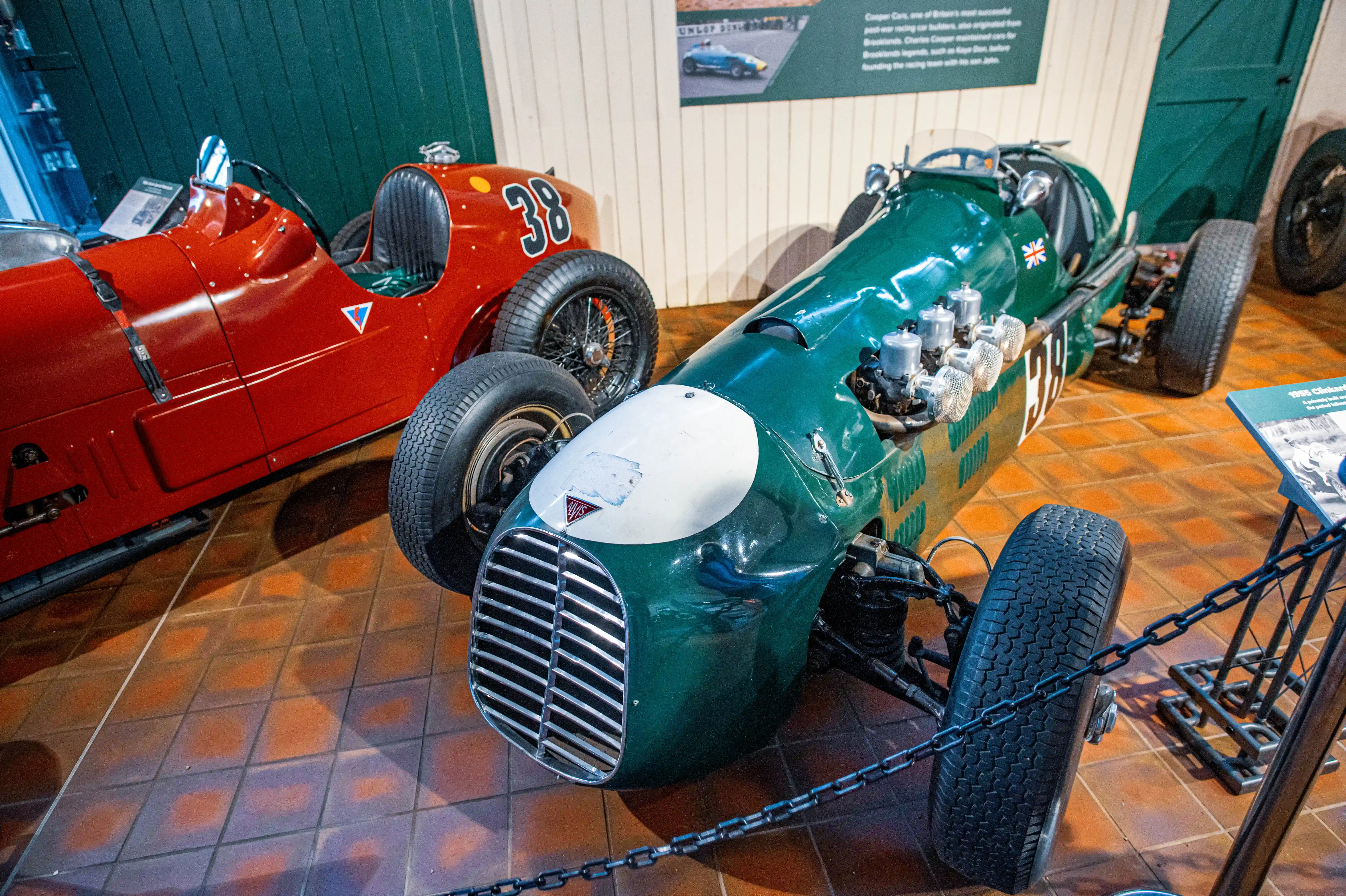 A close-up, eye-level shot shows two vintage open-wheel racing cars in a museum setting. The car in the foreground is predominantly green with a white nose cone, featuring a visible V8 engine setup and a Union Jack flag emblem. The car to the left is bright red with the number '38' on its side. Both cars have classic spoked wheels and thin racing tyres. The floor is tiled with reddish-brown squares.