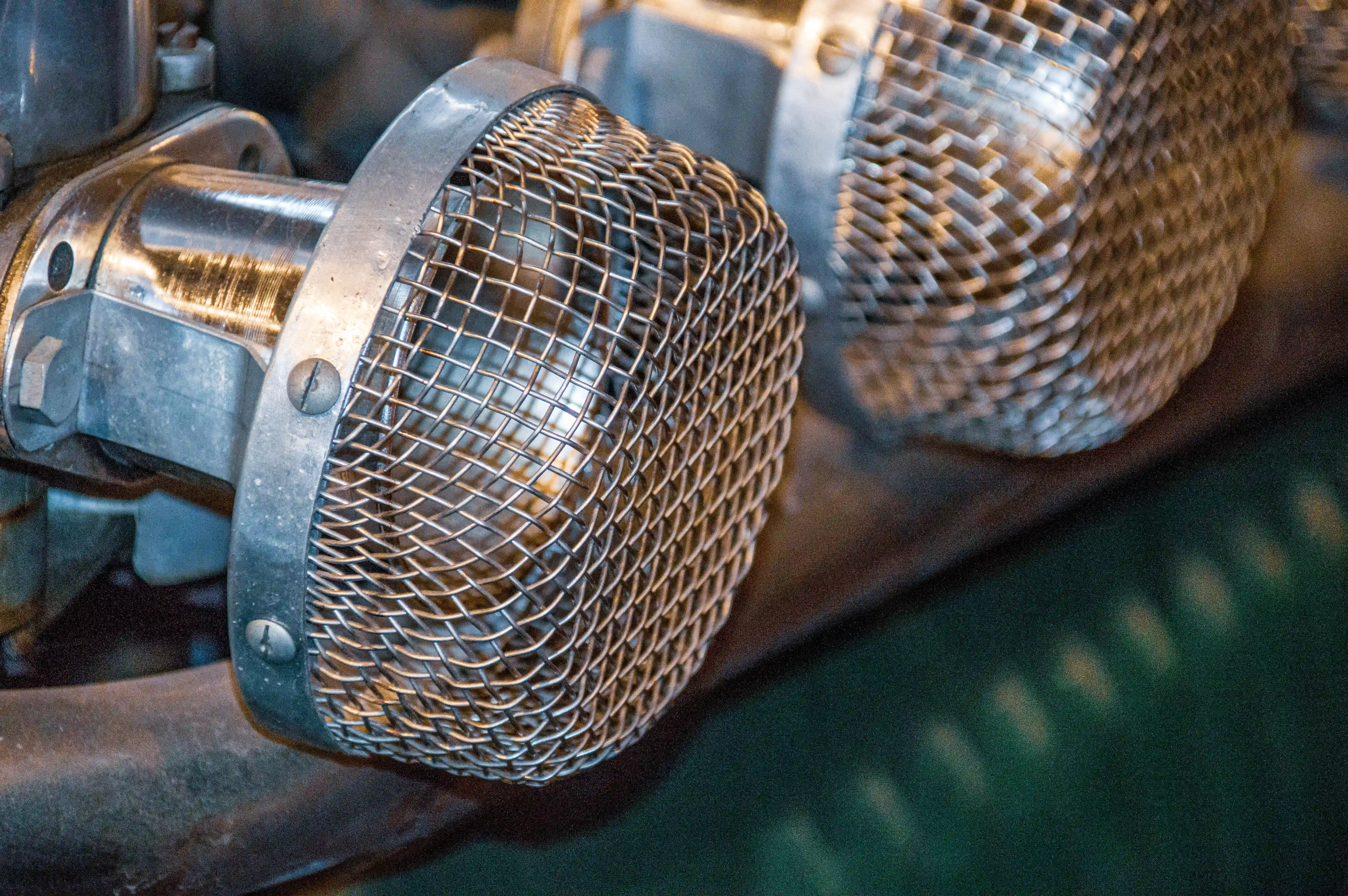 Close-up of two shiny metal air intake filters with fine mesh, attached to a vintage car engine.