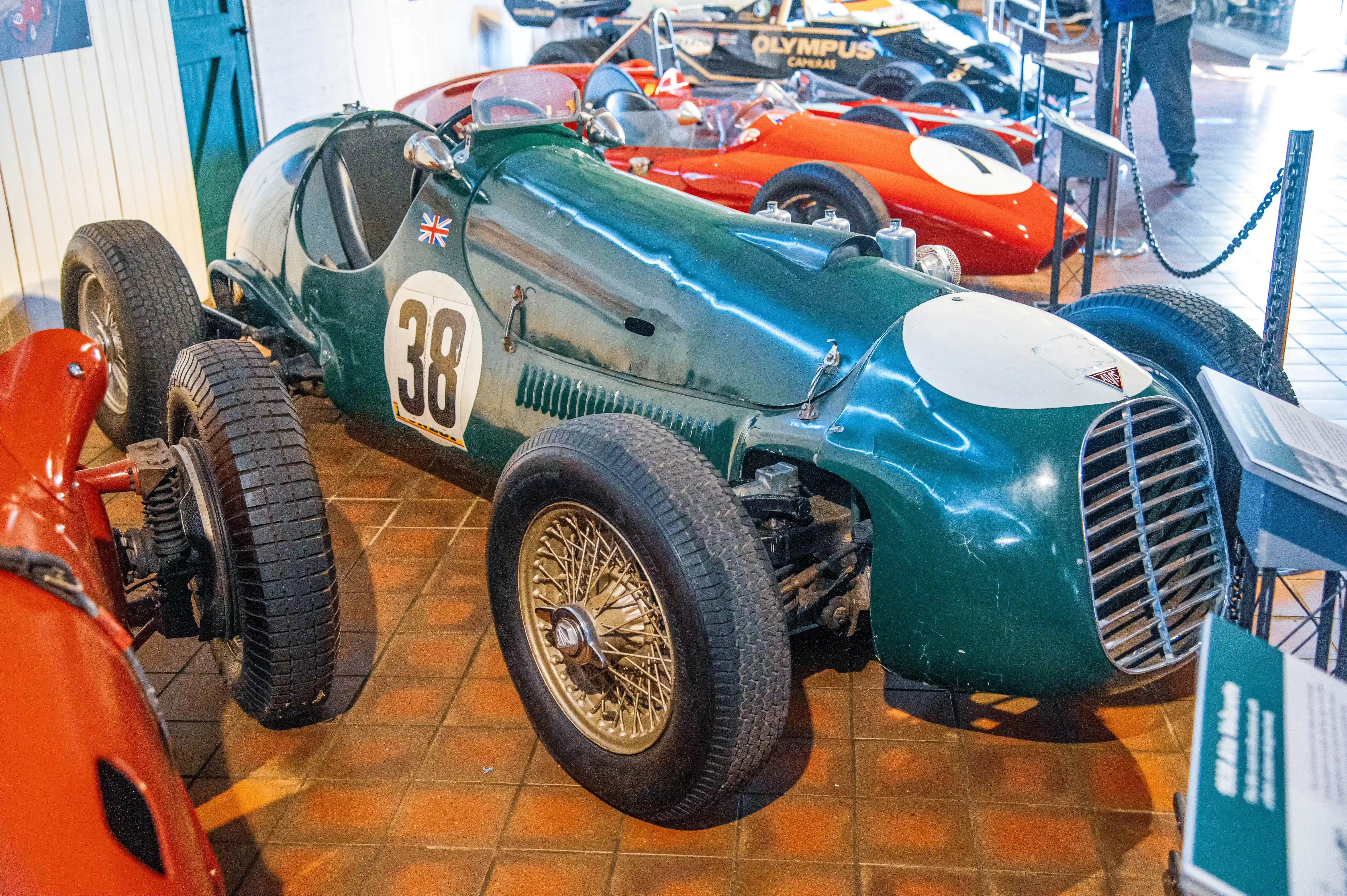 A green vintage racing car with the number 38 and a Union Jack flag is displayed in a museum next to a red racing car. Informational plaques are visible in front of both vehicles.