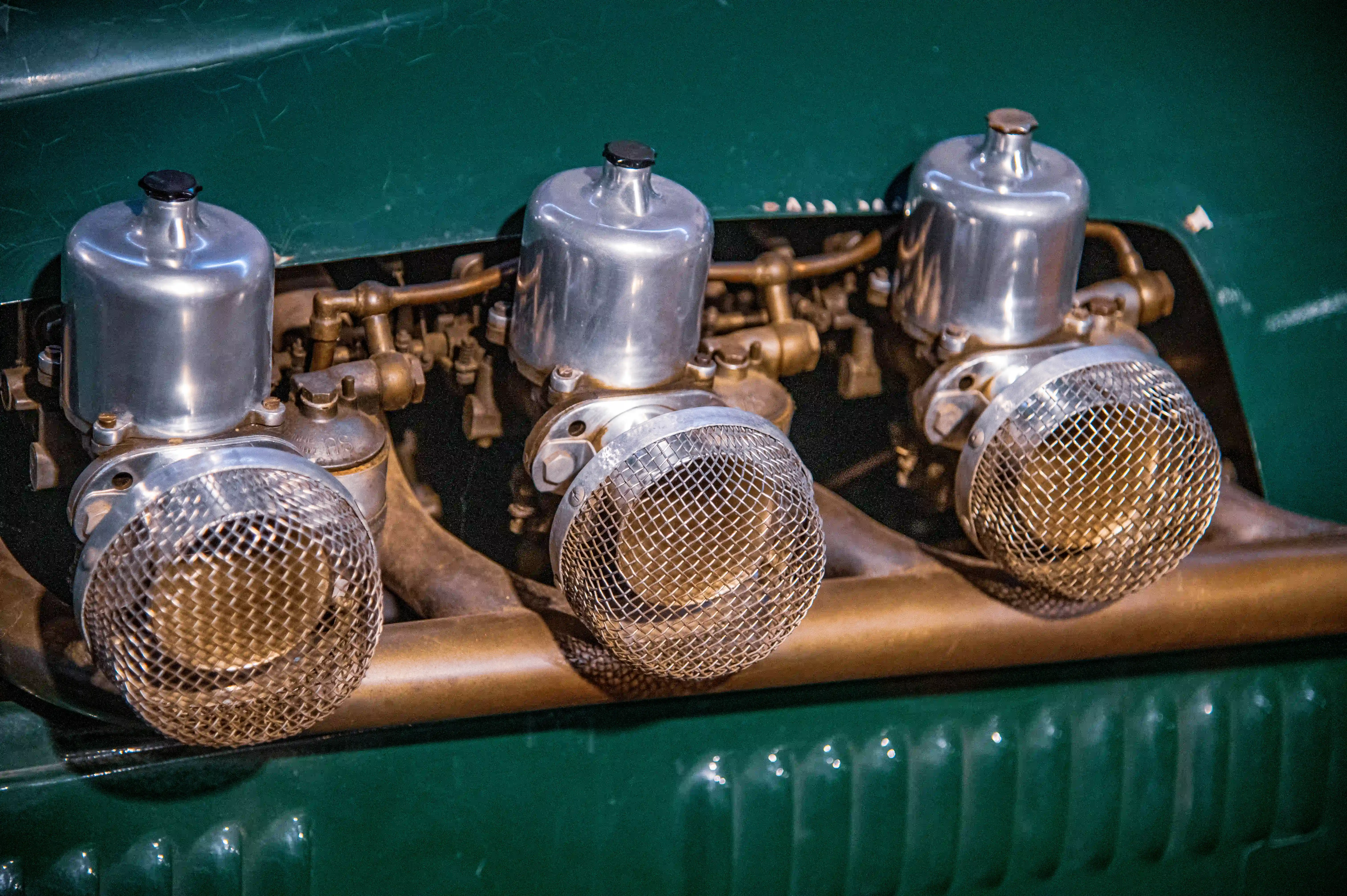Three polished metal carburettors with wire mesh intake filters arranged in a row, mounted on a vintage vehicle with a deep green painted engine bay.