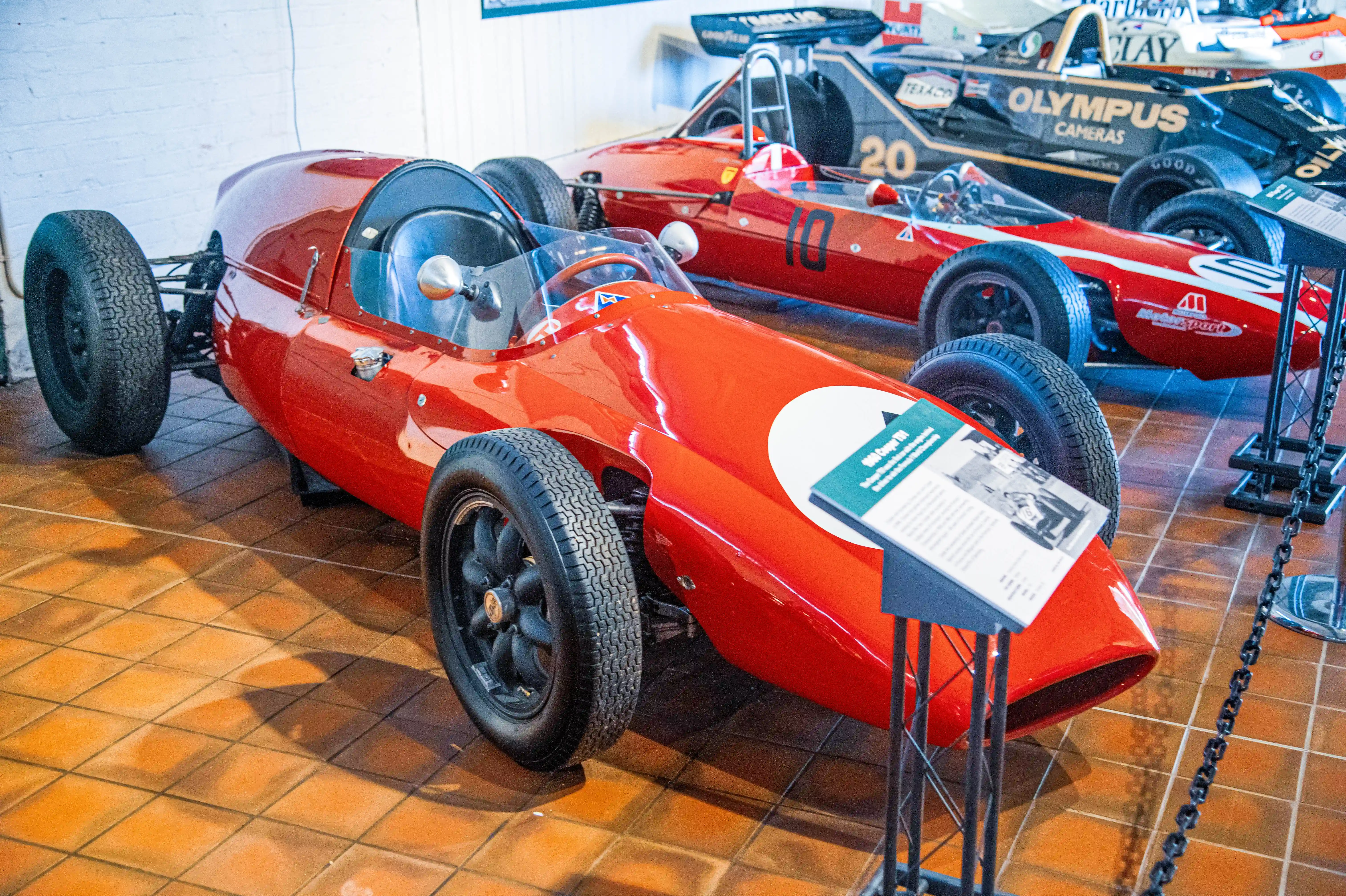 Close-up of a bright red vintage Formula 1 racing car, its sleek bodywork reflecting the light. Another red car with the number '10' and a black and gold car with the number '20' are visible behind it. A descriptive plaque is placed on the front of the red car.