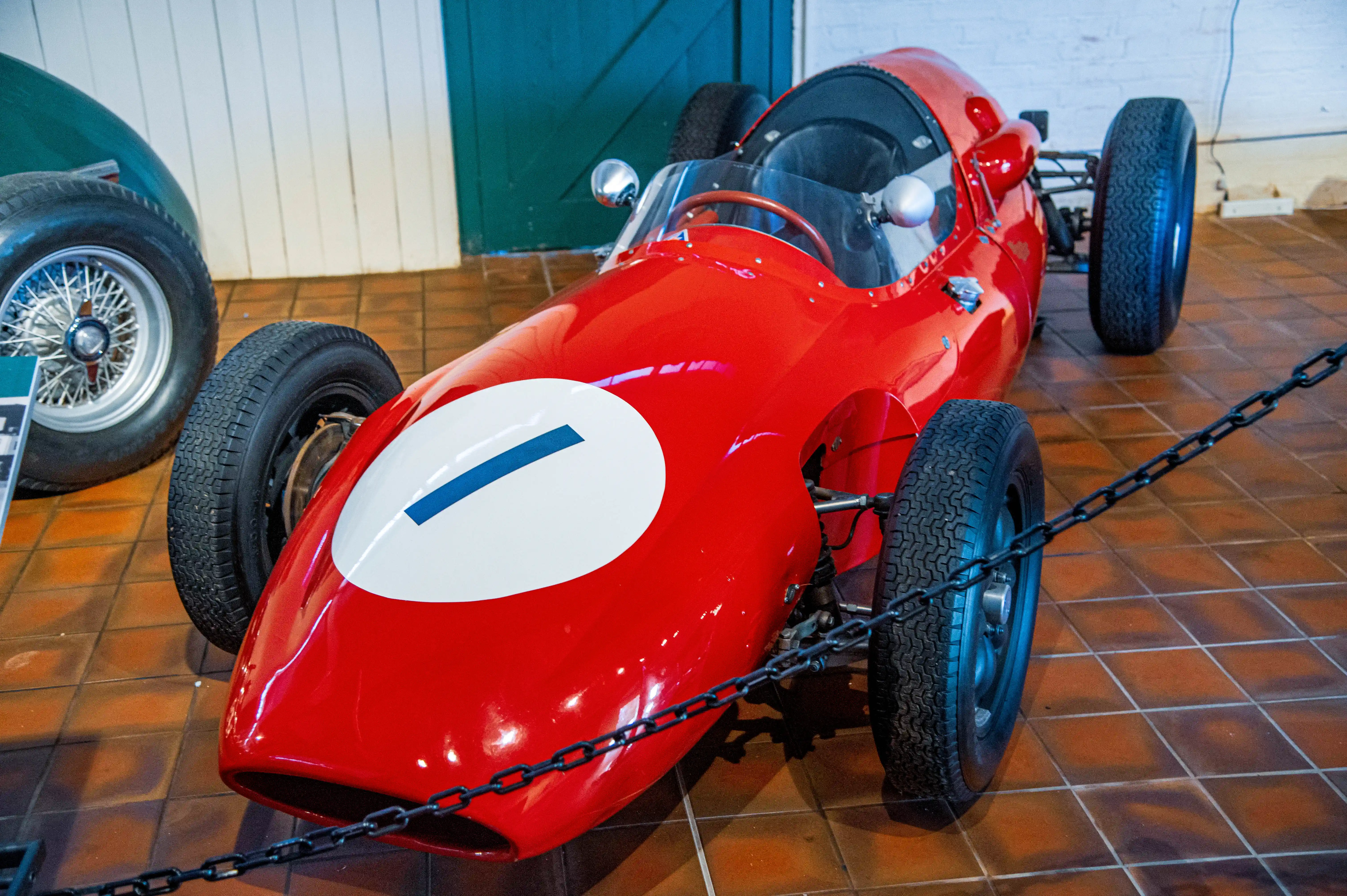 Close-up of a bright red vintage Formula 1 racing car, its sleek bodywork reflecting the light. Another red car with the number '10' and a black and gold car with the number '20' are visible behind it. A descriptive plaque is placed on the front of the red car.