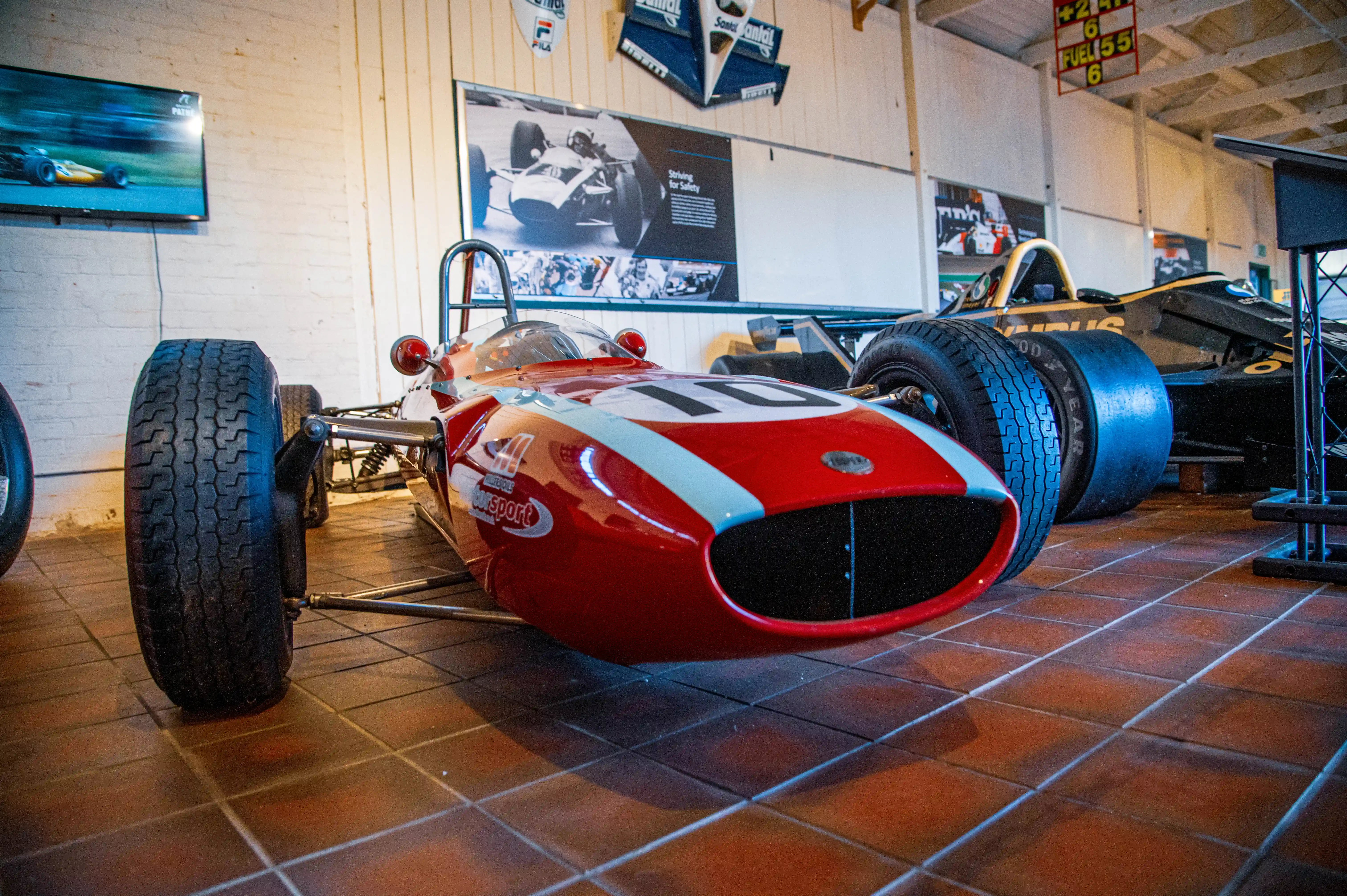 A vintage red and white racing car with number 10 on display, showcasing its aerodynamic design and large wheels. Another black racing car is visible in the background, with memorabilia on the wall.