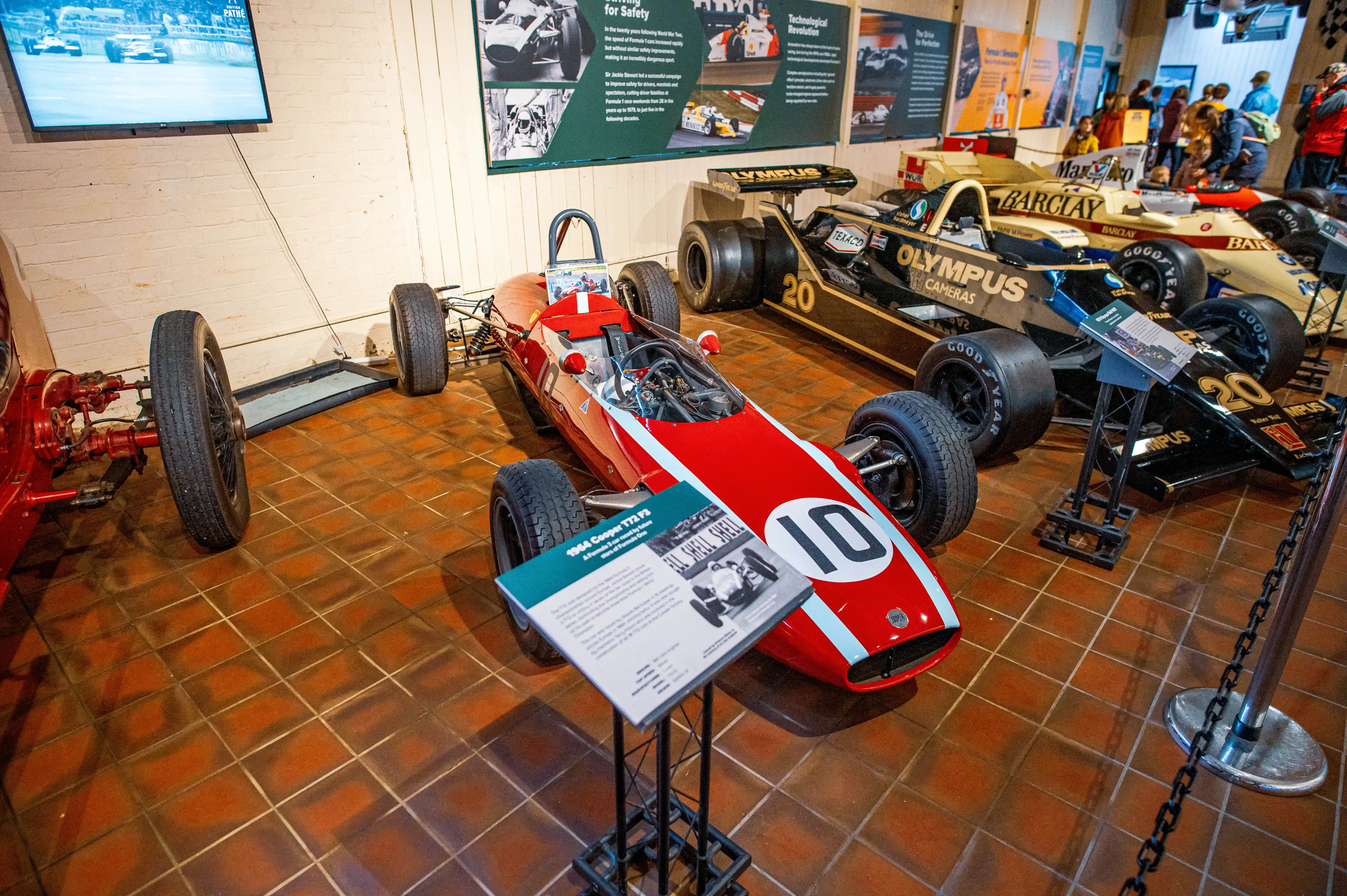 A vintage red and white racing car with number 10 on display, showcasing its aerodynamic design and large wheels. Another black racing car is visible in the background, with memorabilia on the wall.