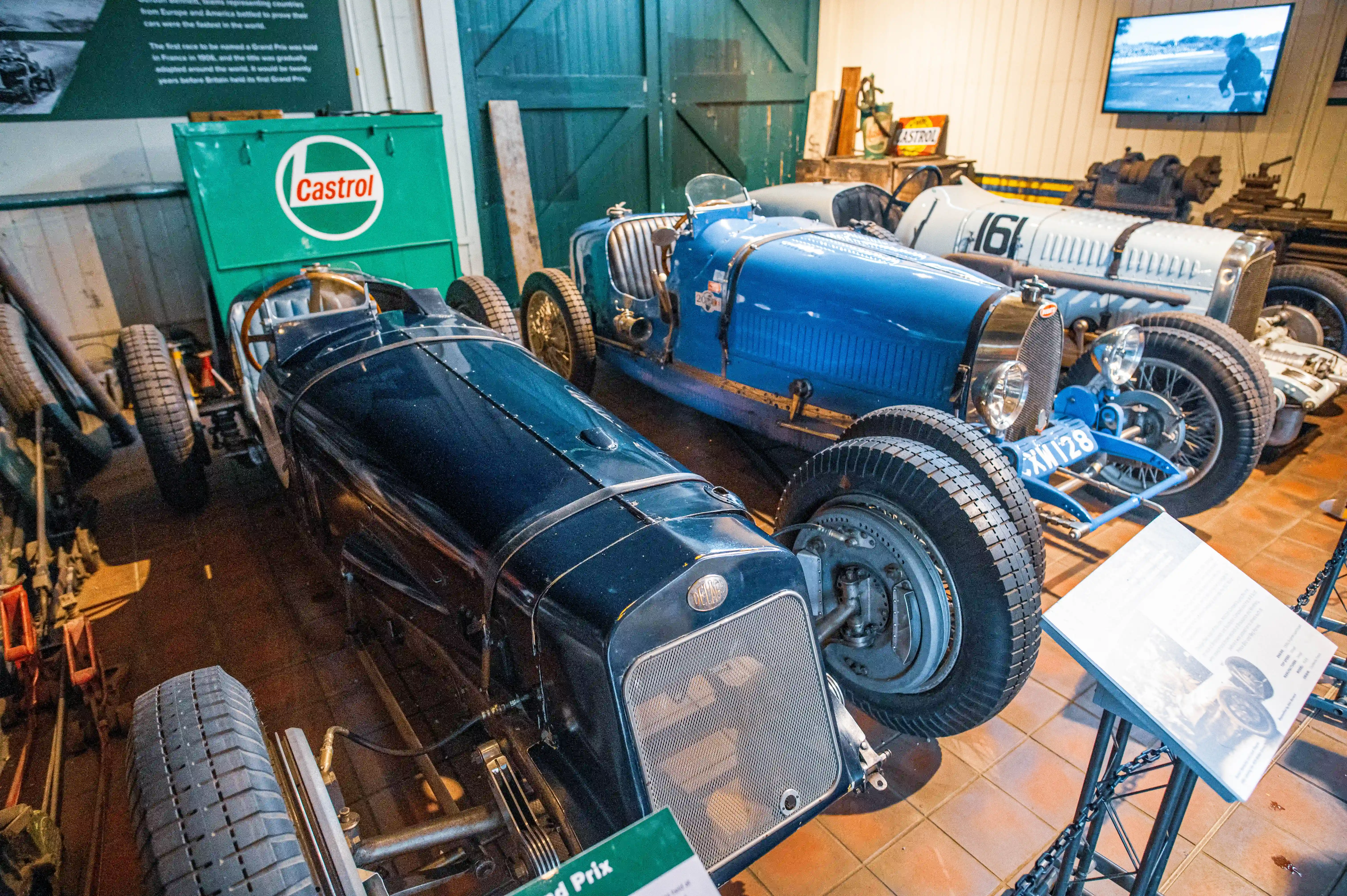 Two vintage Grand Prix racing cars parked in a museum. The car in the foreground is dark blue and the car behind it is bright blue. A green Castrol oil can is visible to the left of the cars.