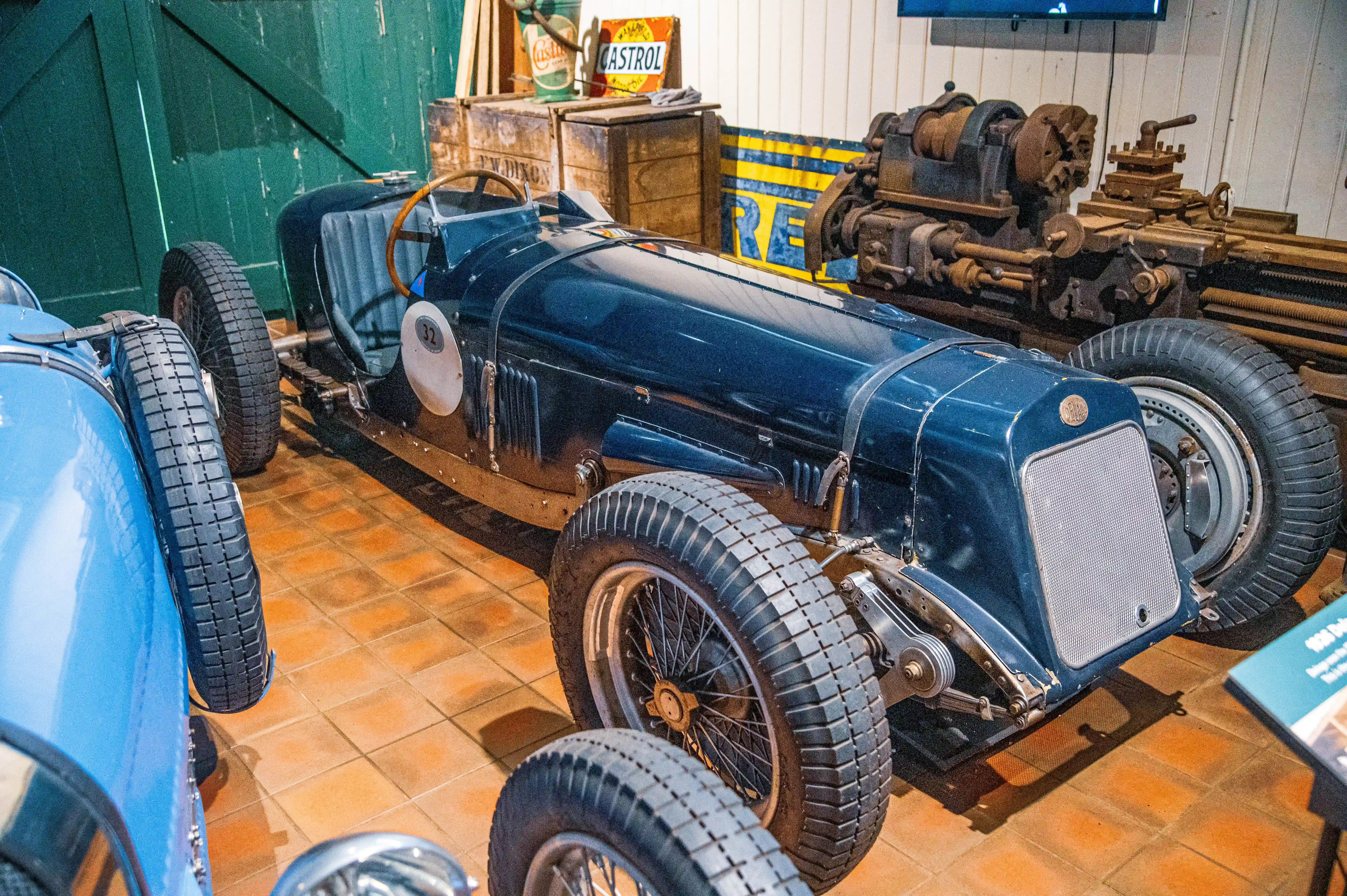 A vintage dark blue racing car with spoked wheels and ribbed tyres is parked indoors. Information boards and vintage machinery are present in the background.