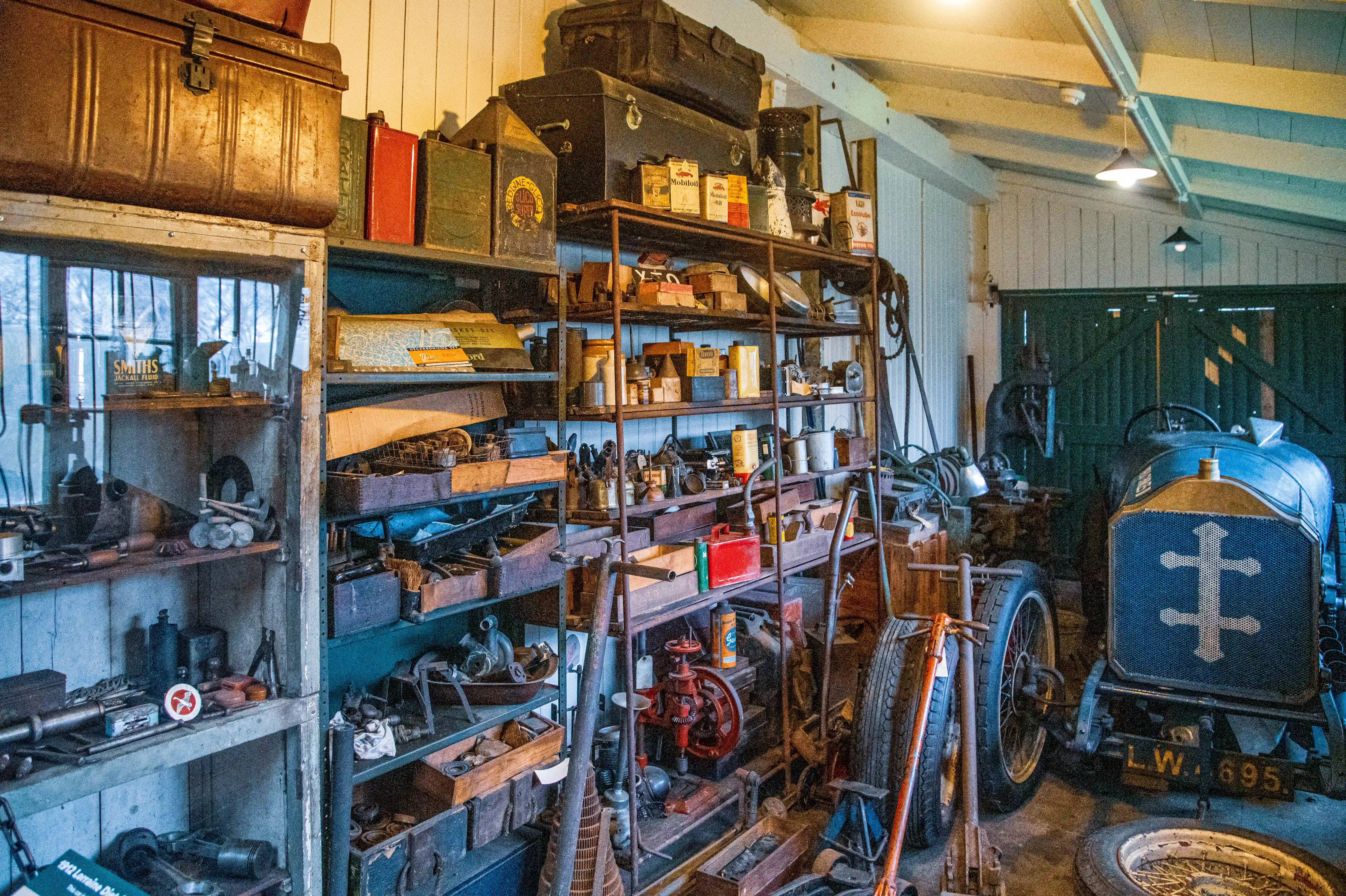  Interior of a vintage garage workshop with shelves of old tools, oil cans, and spare parts. A blue vintage car is parked on the right.