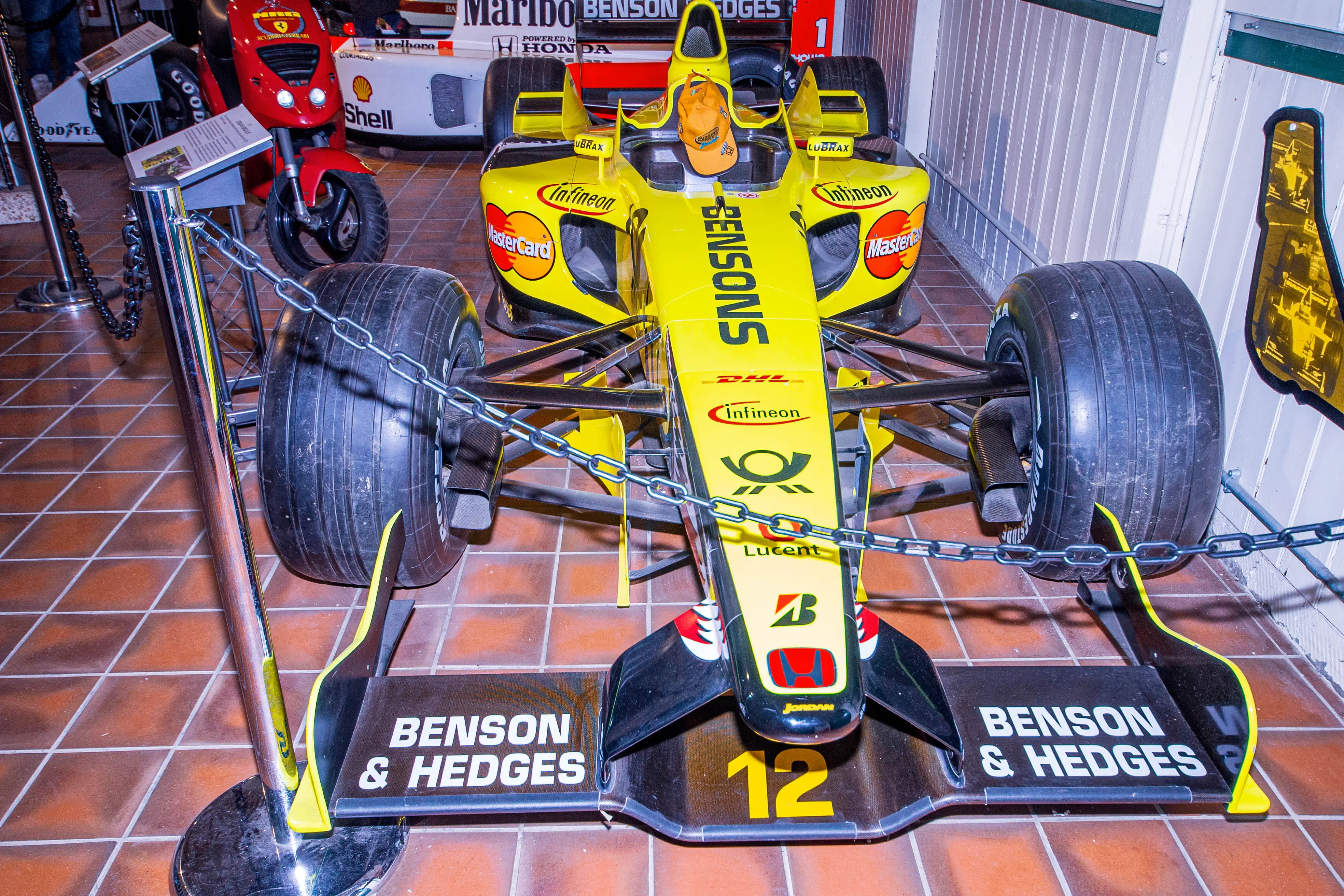 A close-up, front-on view of a yellow and black Jordan Formula 1 racing car, number 12, with 'BENSON & HEDGES' branding on the front wing, on display.