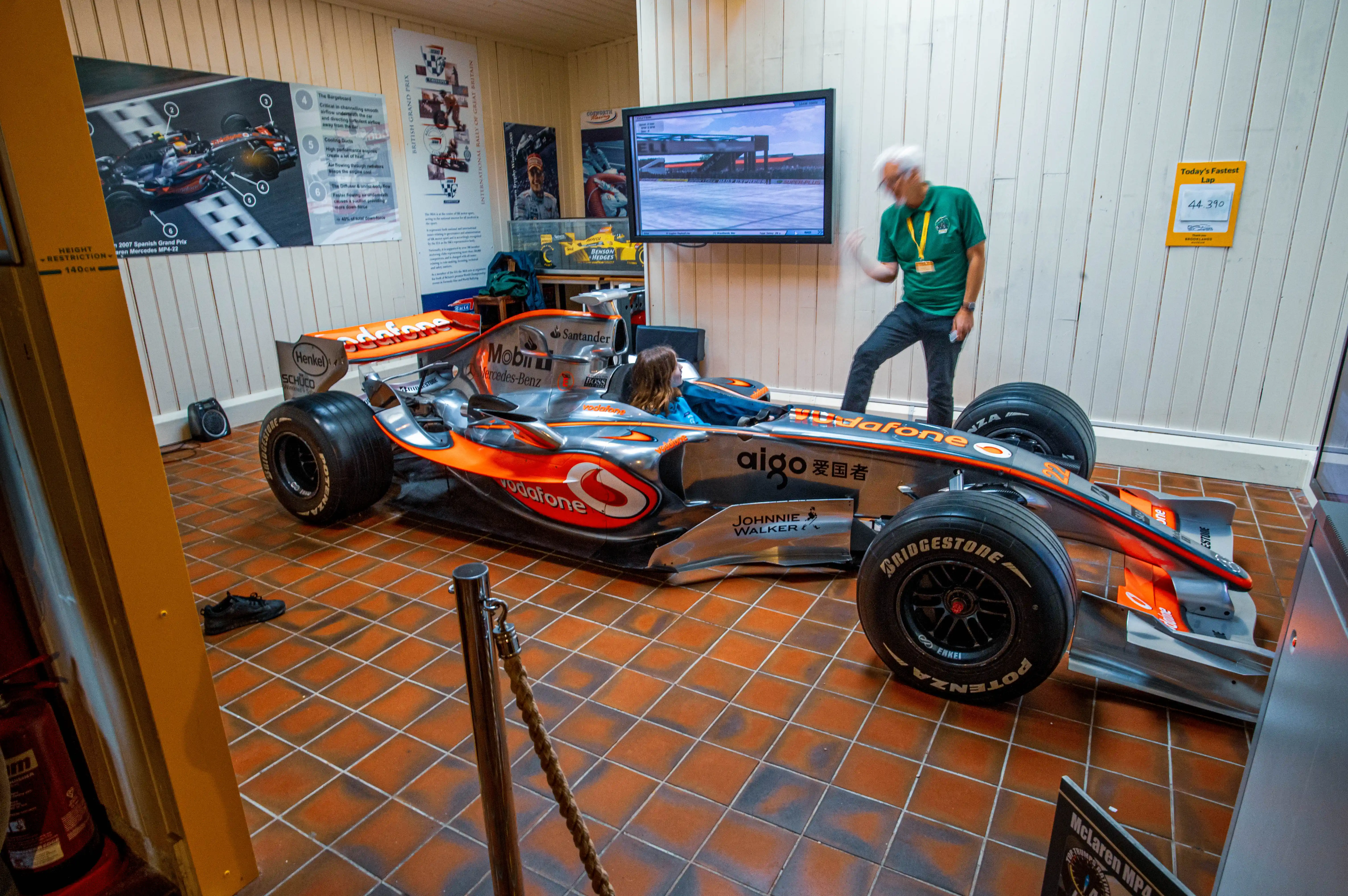 A silver and orange McLaren Formula 1 racing car is exhibited in a museum. A man in a green shirt stands beside it, gesturing towards a television screen displaying a racing circuit. A young girl is sitting inside the car's cockpit. The floor is tiled with terracotta squares, and various posters and information boards are visible on the walls.