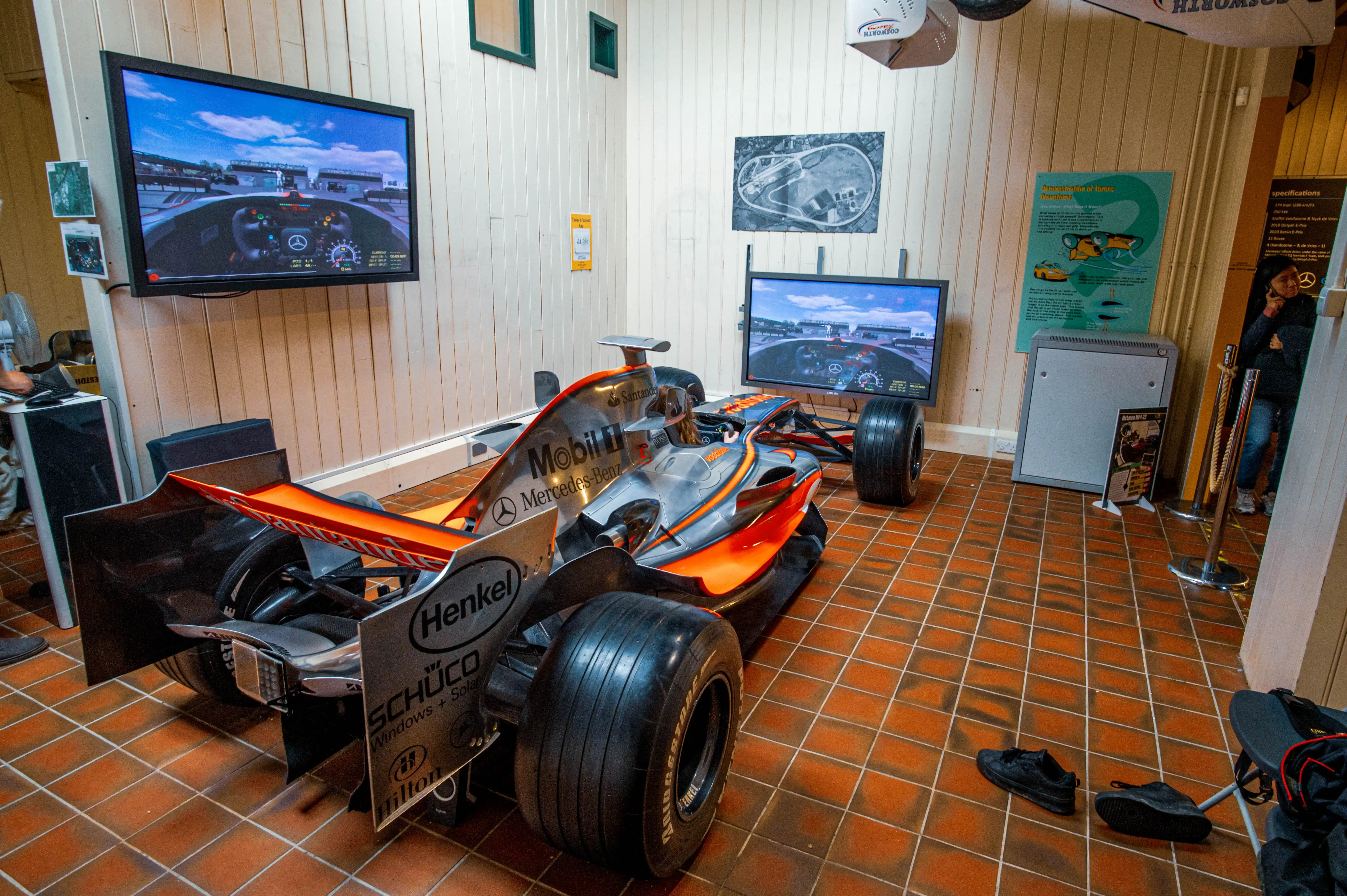 A silver and orange McLaren Formula 1 racing car is exhibited in a museum. A man in a green shirt stands beside it, gesturing towards a television screen displaying a racing circuit. A young girl is sitting inside the car's cockpit. The floor is tiled with terracotta squares, and various posters and information boards are visible on the walls.