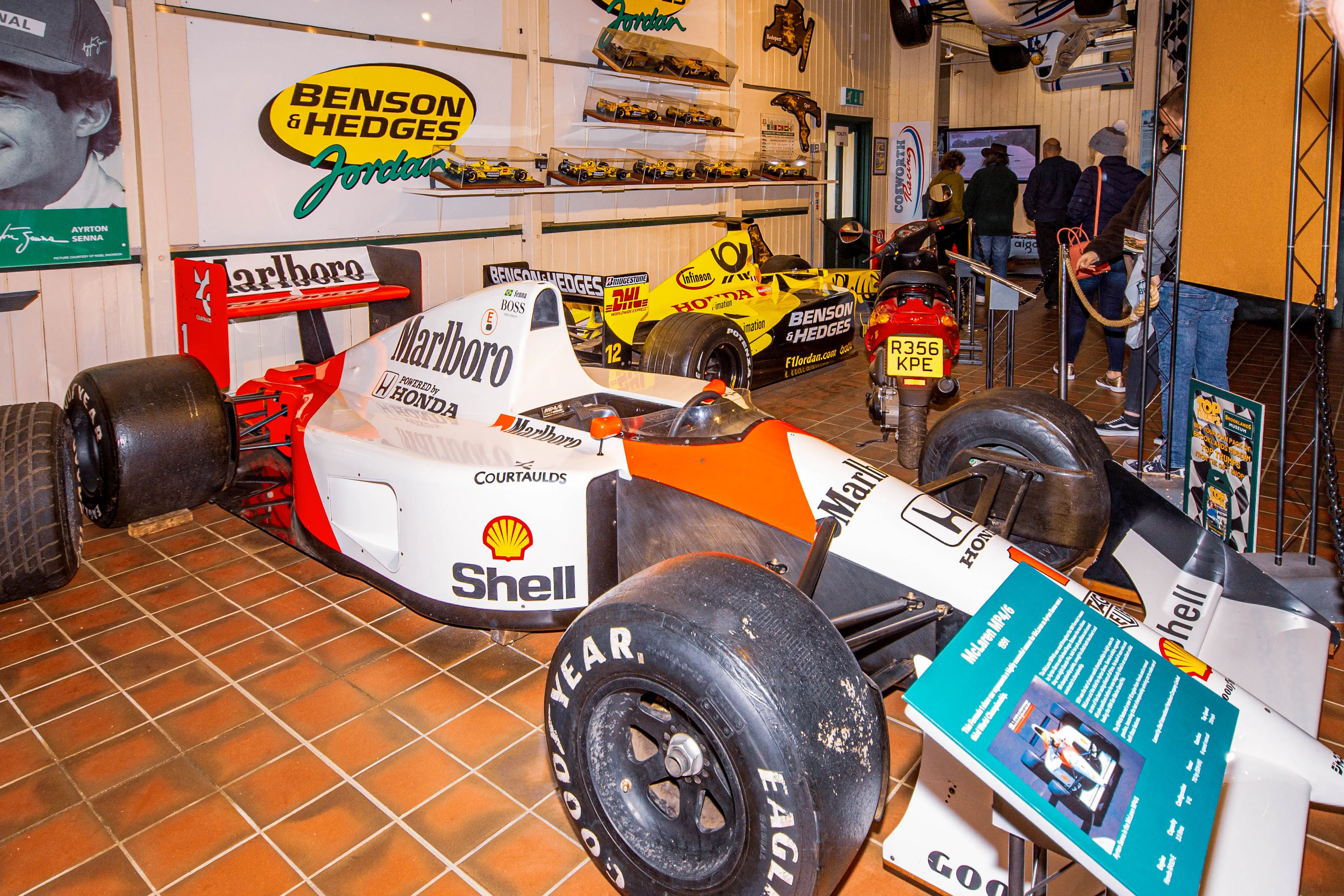 A white and red Formula 1 car, identified as a McLaren MP4/6, with prominent Marlboro and Shell logos, is displayed on a tiled floor in a museum. A yellow Jordan Formula 1 car is visible in the background, as are other exhibits and visitors.