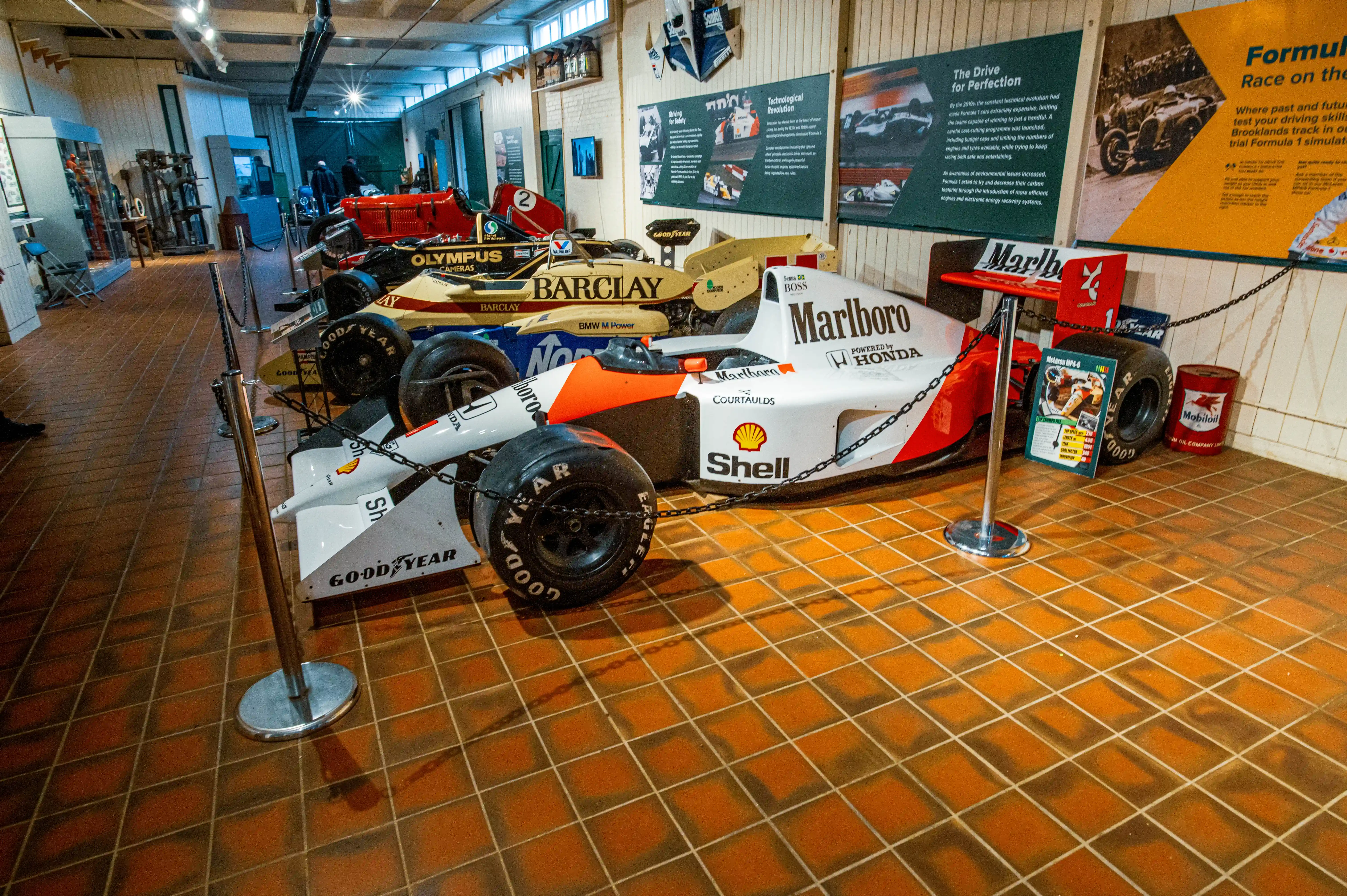 A white and red Formula 1 car, identified as a McLaren MP4/6, with prominent Marlboro and Shell logos, is displayed on a tiled floor in a museum. A yellow Jordan Formula 1 car is visible in the background, as are other exhibits and visitors.