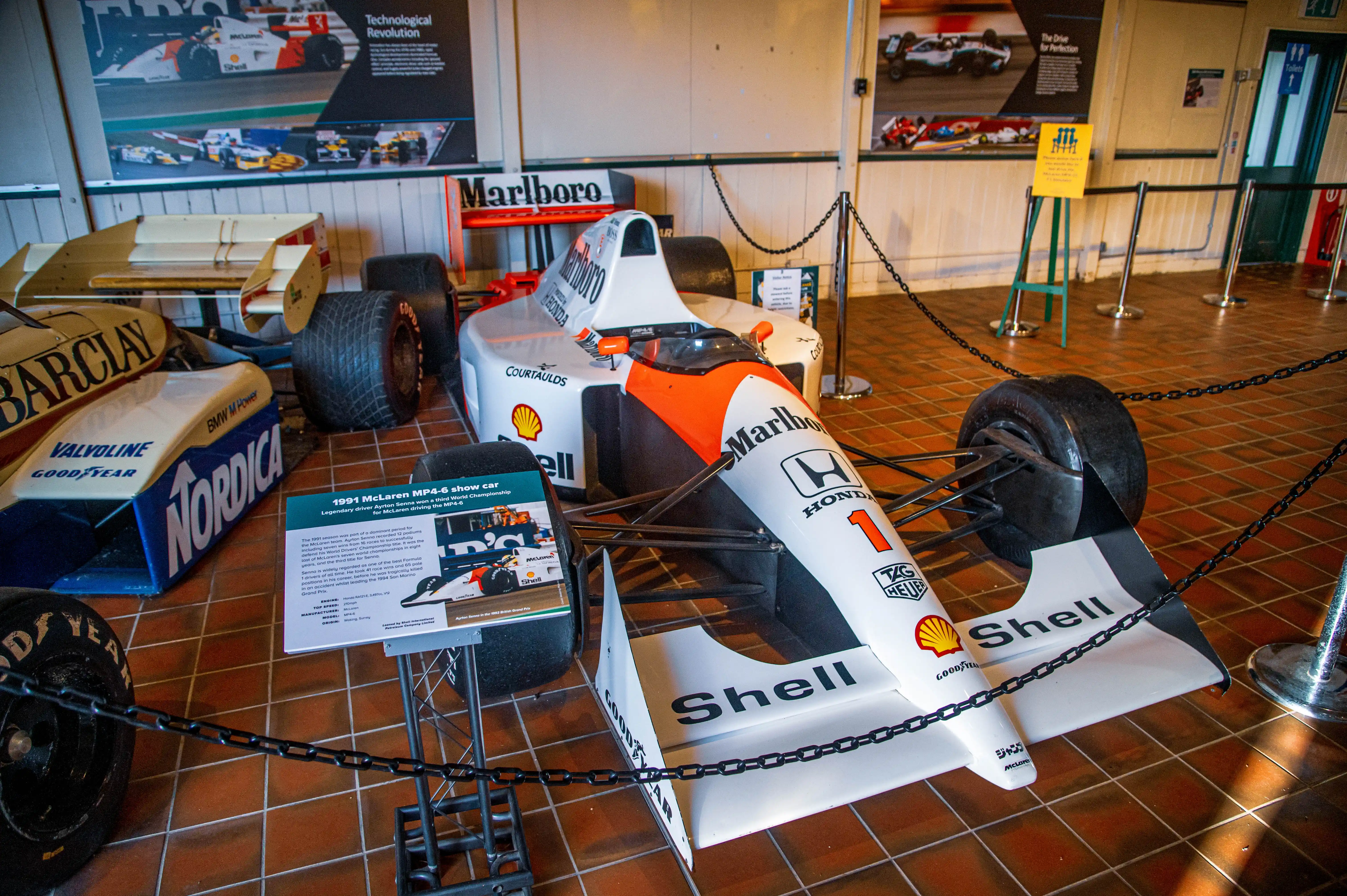 A white and orange McLaren MP4-6 Formula 1 racing car with the number 1 on the front wing is parked on a tiled floor, with a descriptive plaque in front of it. Other racing car parts and posters are visible in the background.