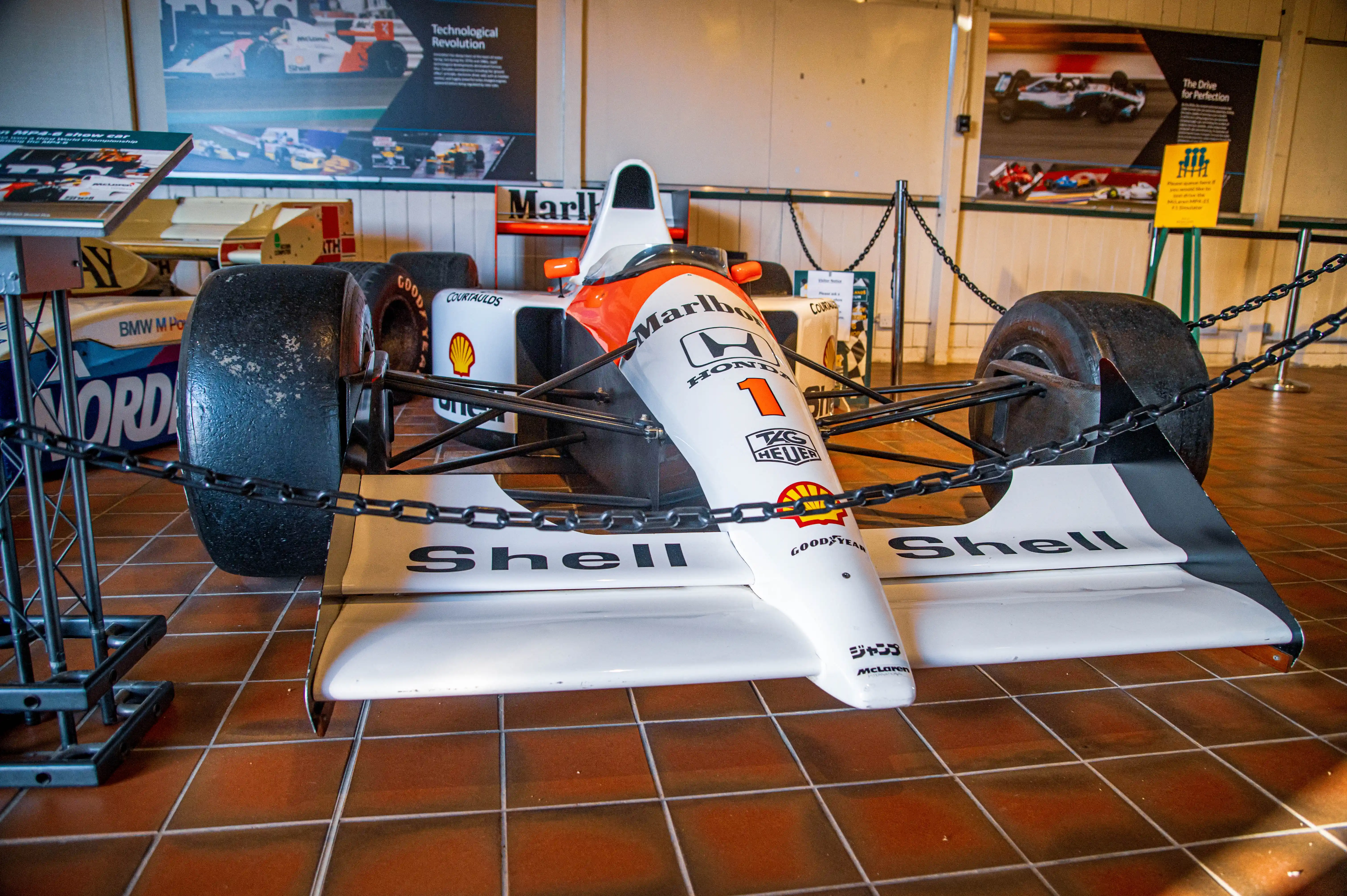 A white and orange McLaren MP4-6 Formula 1 racing car with the number 1 on the front wing is parked on a tiled floor, with a descriptive plaque in front of it. Other racing car parts and posters are visible in the background.