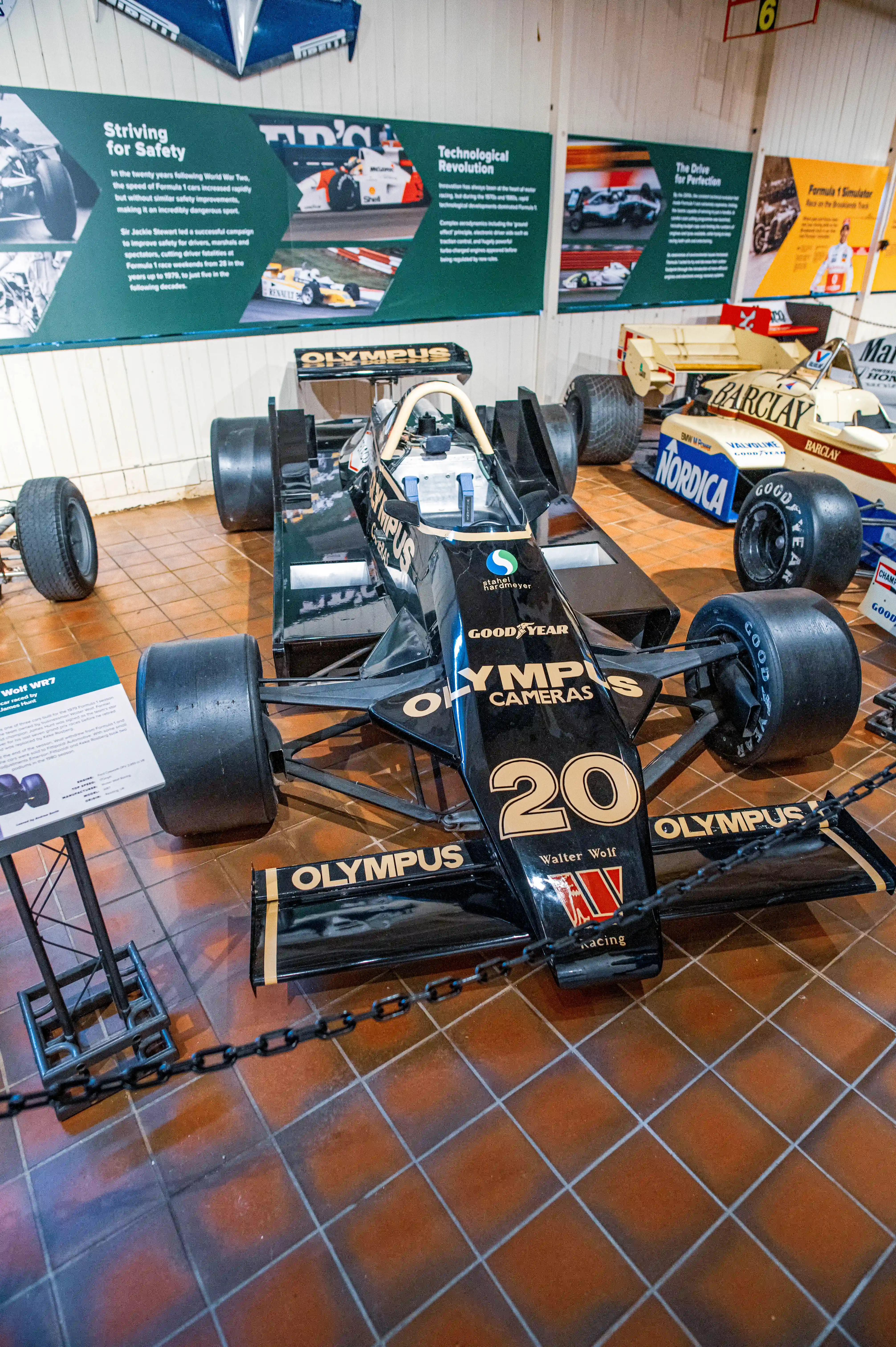 A black and gold Formula 1 racing car from the 1970s, identified as a Walter Wolf WR7, is parked indoors on a tiled floor. The car is adorned with sponsor logos. In the background, exhibition panels detail Formula 1's history and technological evolution.
