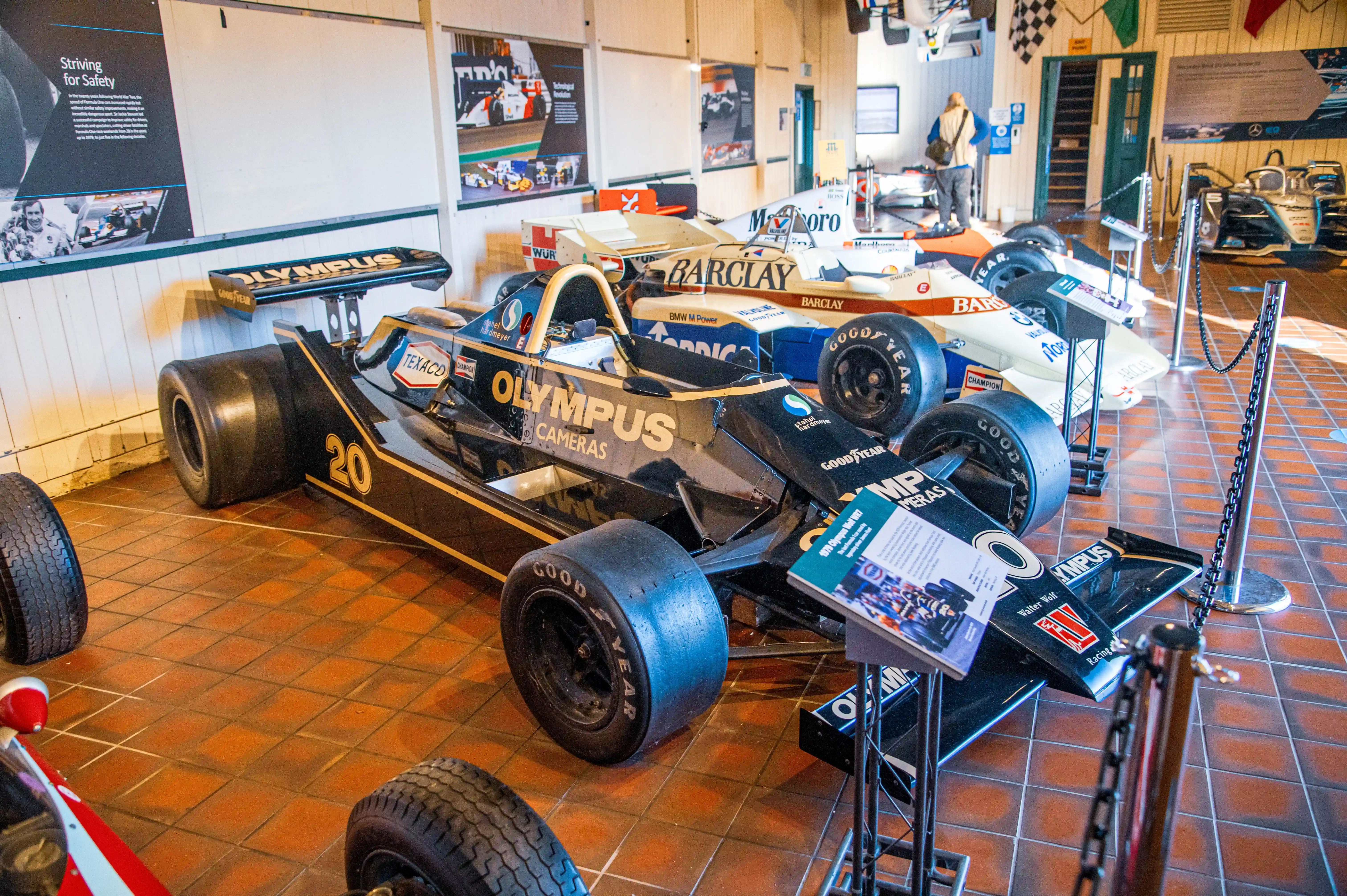A black and gold Formula 1 racing car from the 1970s, identified as a Walter Wolf WR7, is parked indoors on a tiled floor. The car is adorned with sponsor logos. In the background, exhibition panels detail Formula 1's history and technological evolution.